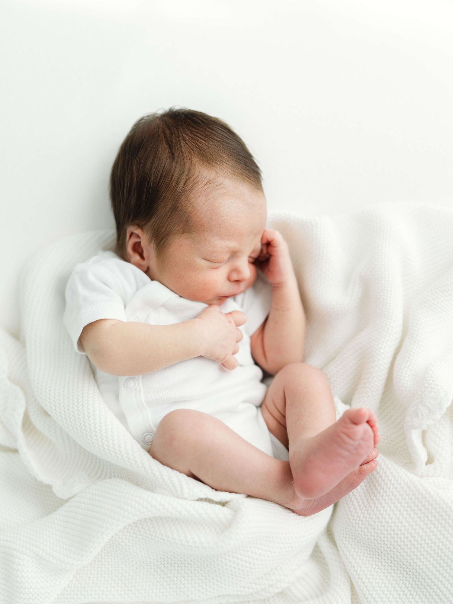 A peaceful newborn baby curled on its side, sleeping on a white blanket in a white onesie while being cared for by nannies in Raleigh.