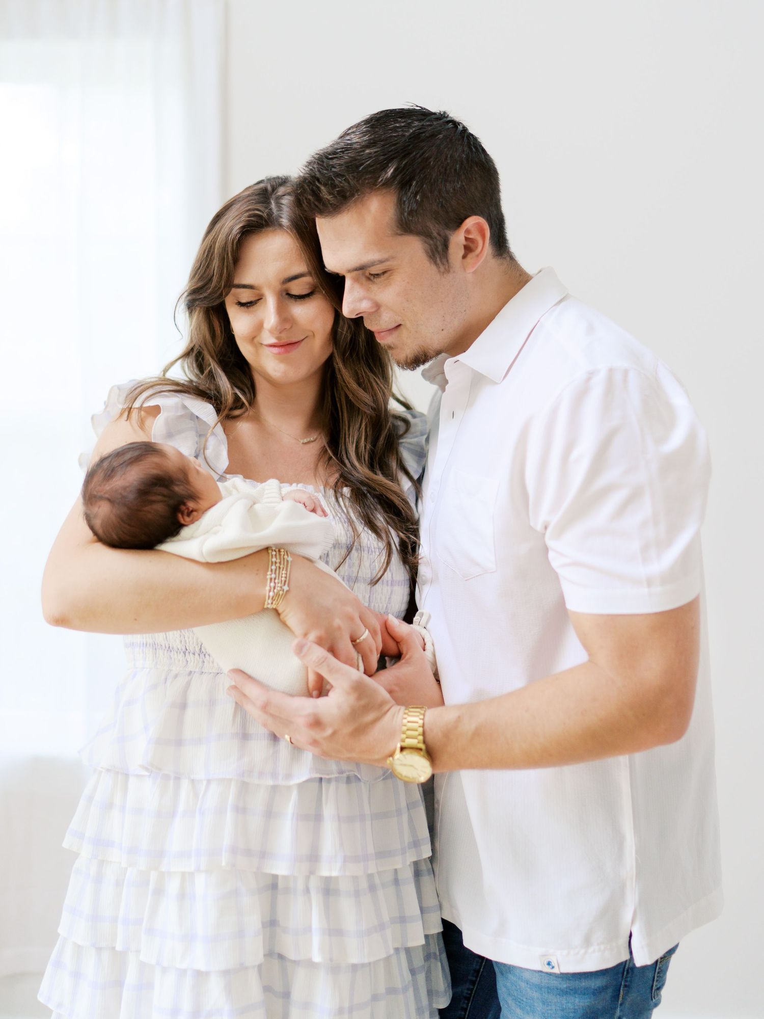New parents in white outfits tenderly hold their sleeping newborn baby during a bright studio photo session.