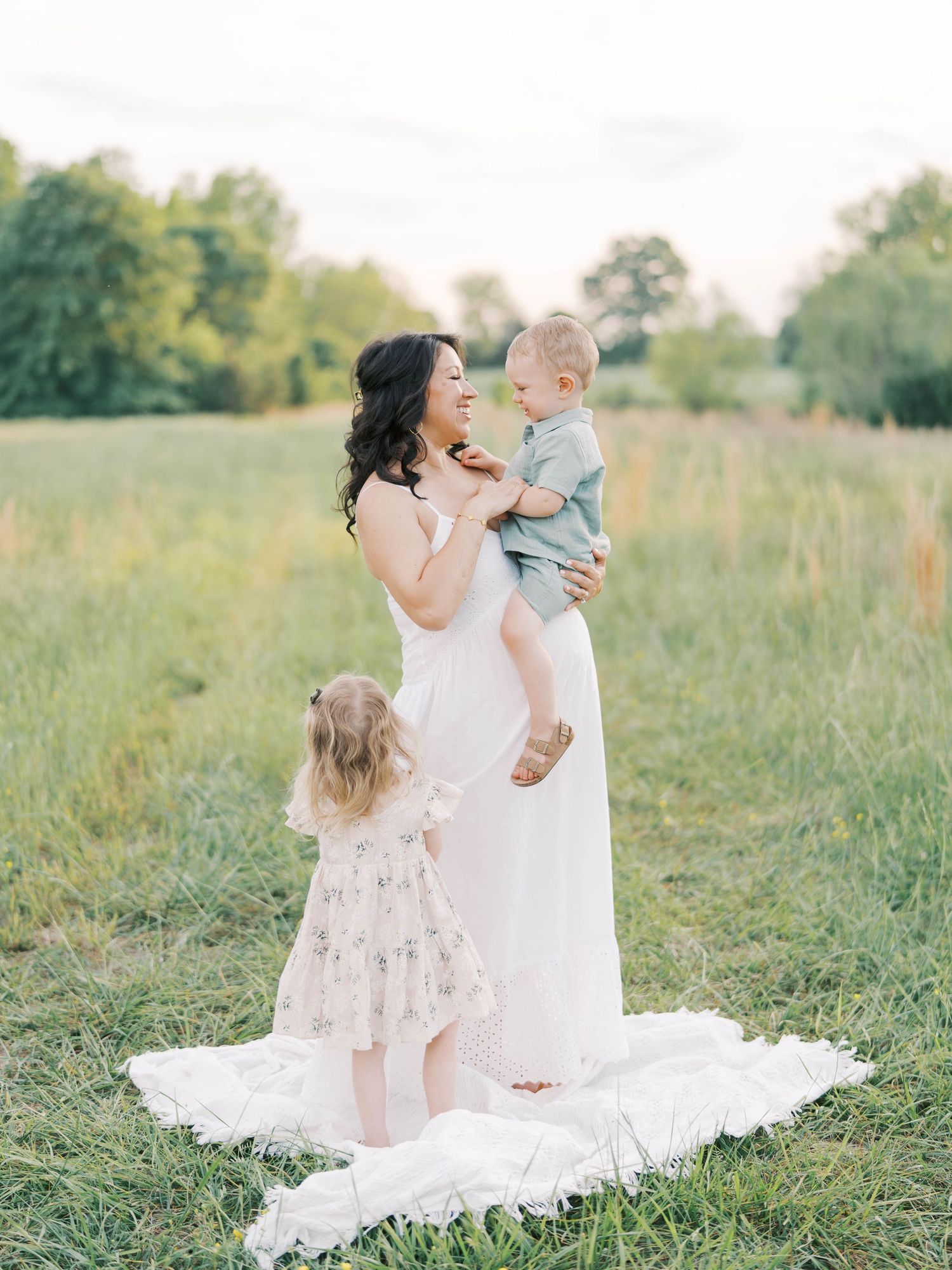 A mother in a white dress holds her baby while her toddler stands beside her in a field before celebrating at baby shower venues in Raleigh.