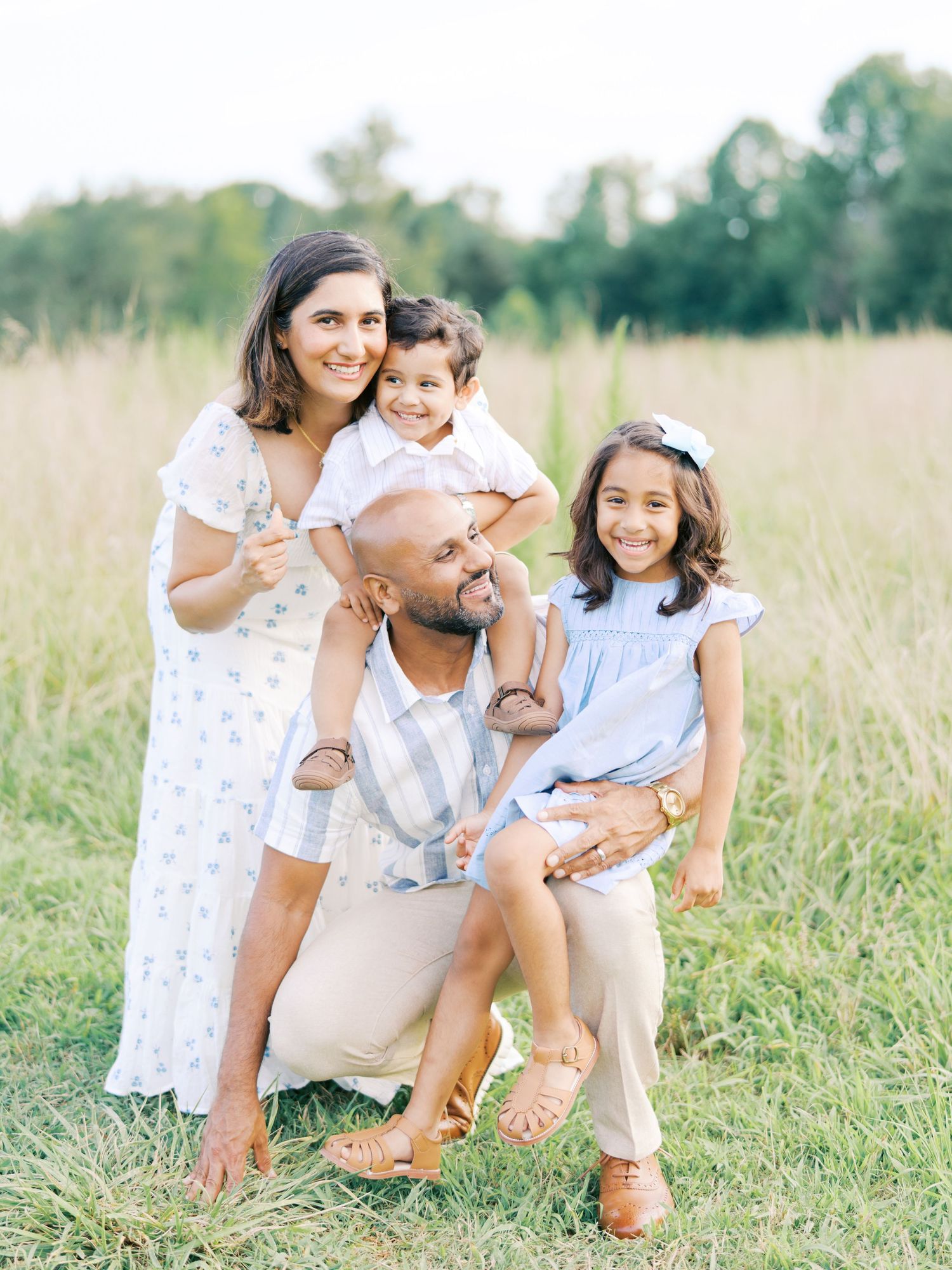 A family sits together in a grassy field wearing coordinated white and blue outfits for a casual outdoor photoshoot.