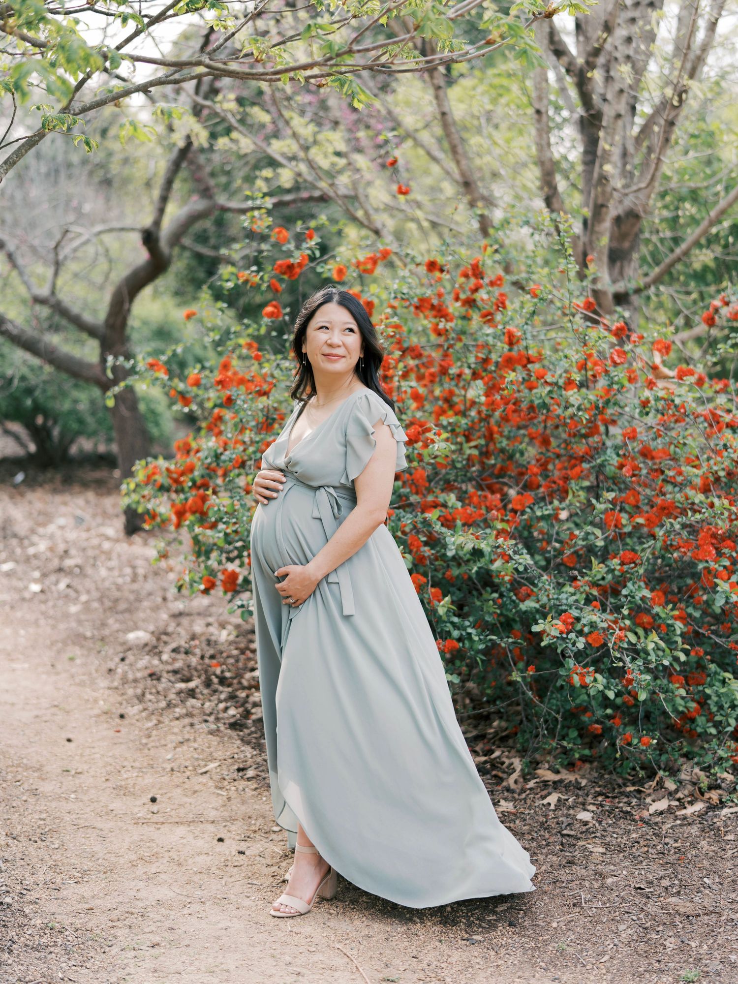 Pregnant woman in blue dress poses by vibrant orange flowering bush on nature trail.