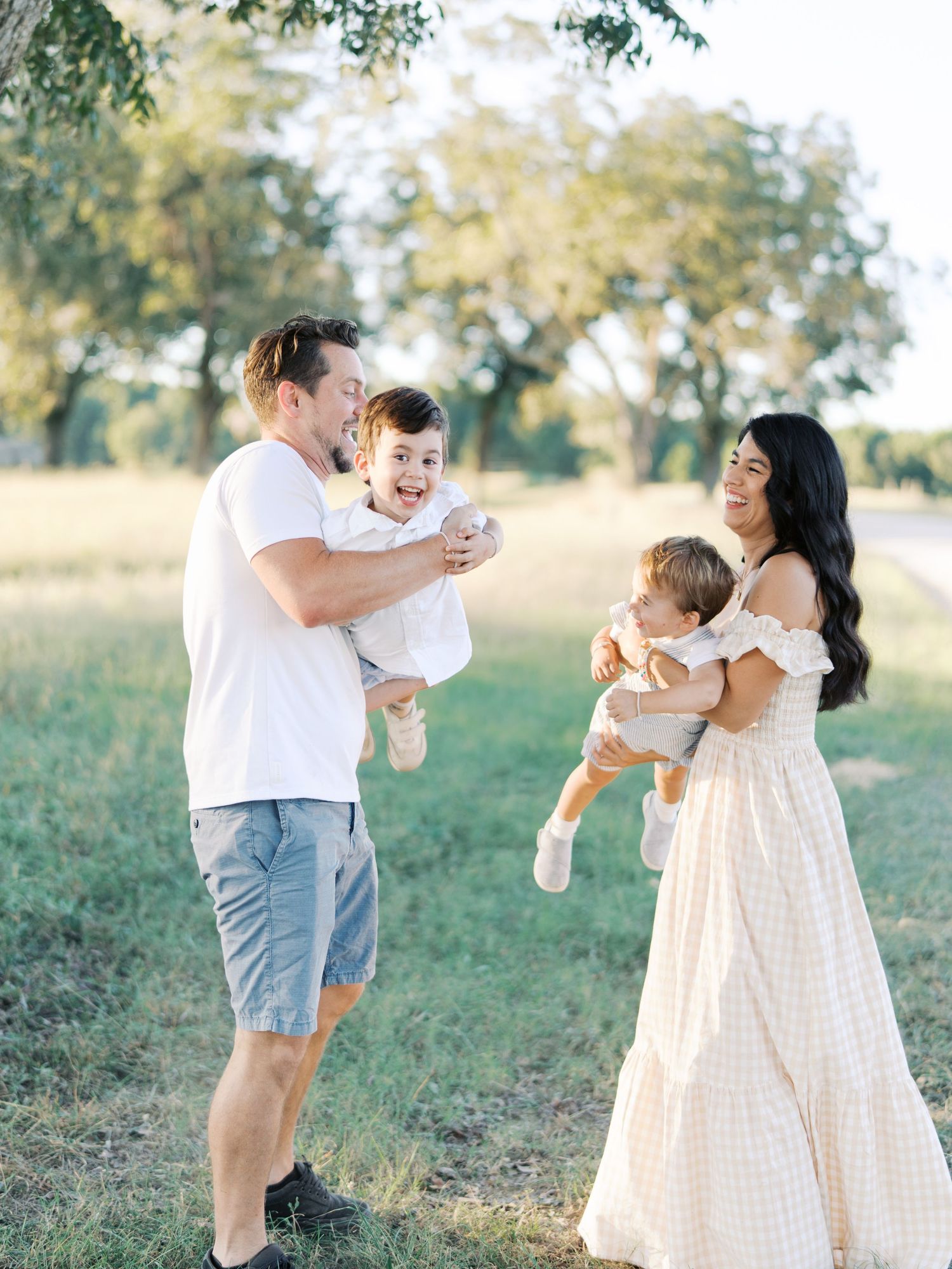 A family wearing white and beige outfits plays and laughs together in a sunny outdoor field during a photo session.