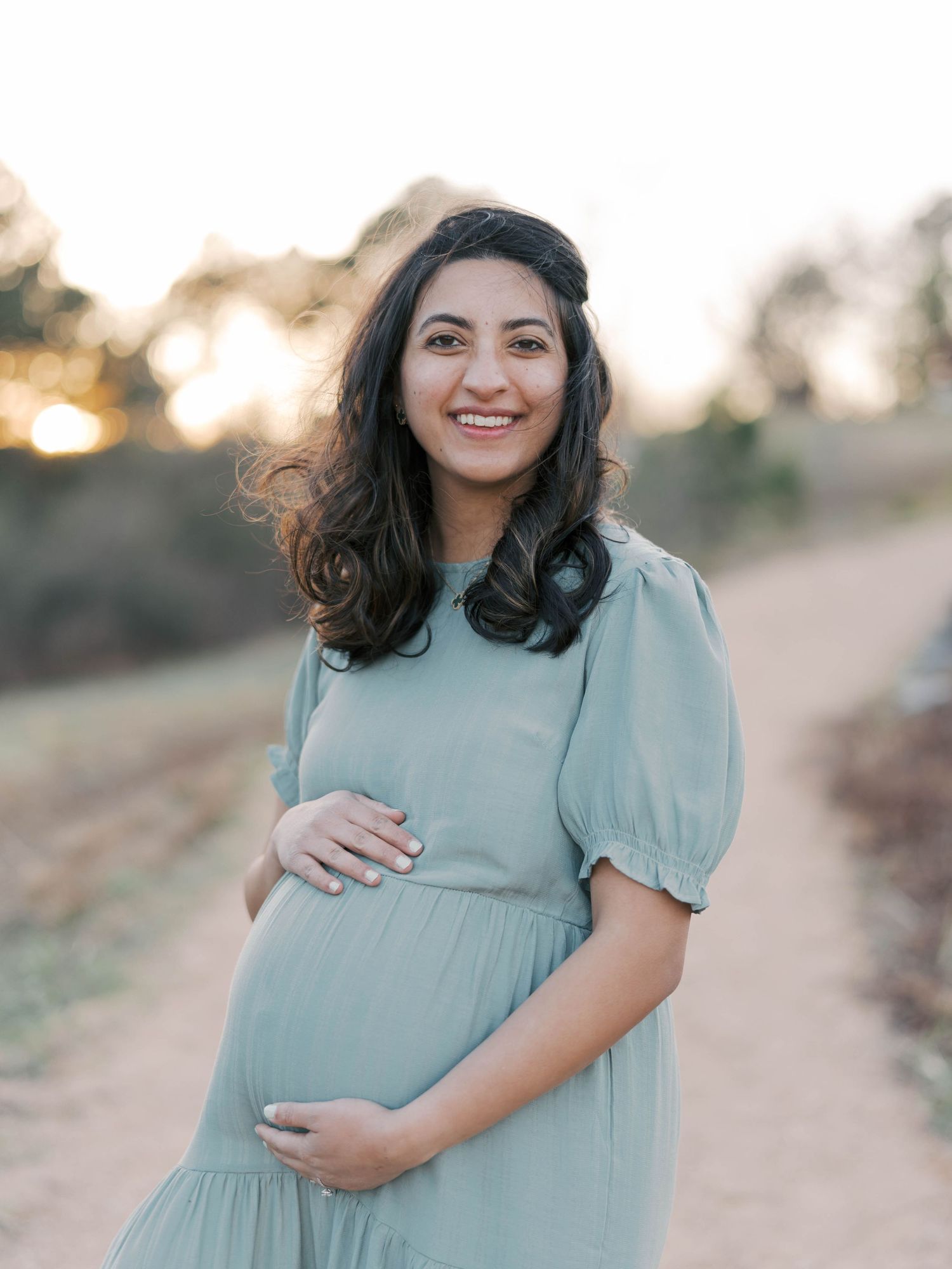 Pregnant woman in blue dress smiling while touching belly on outdoor path at sunset.