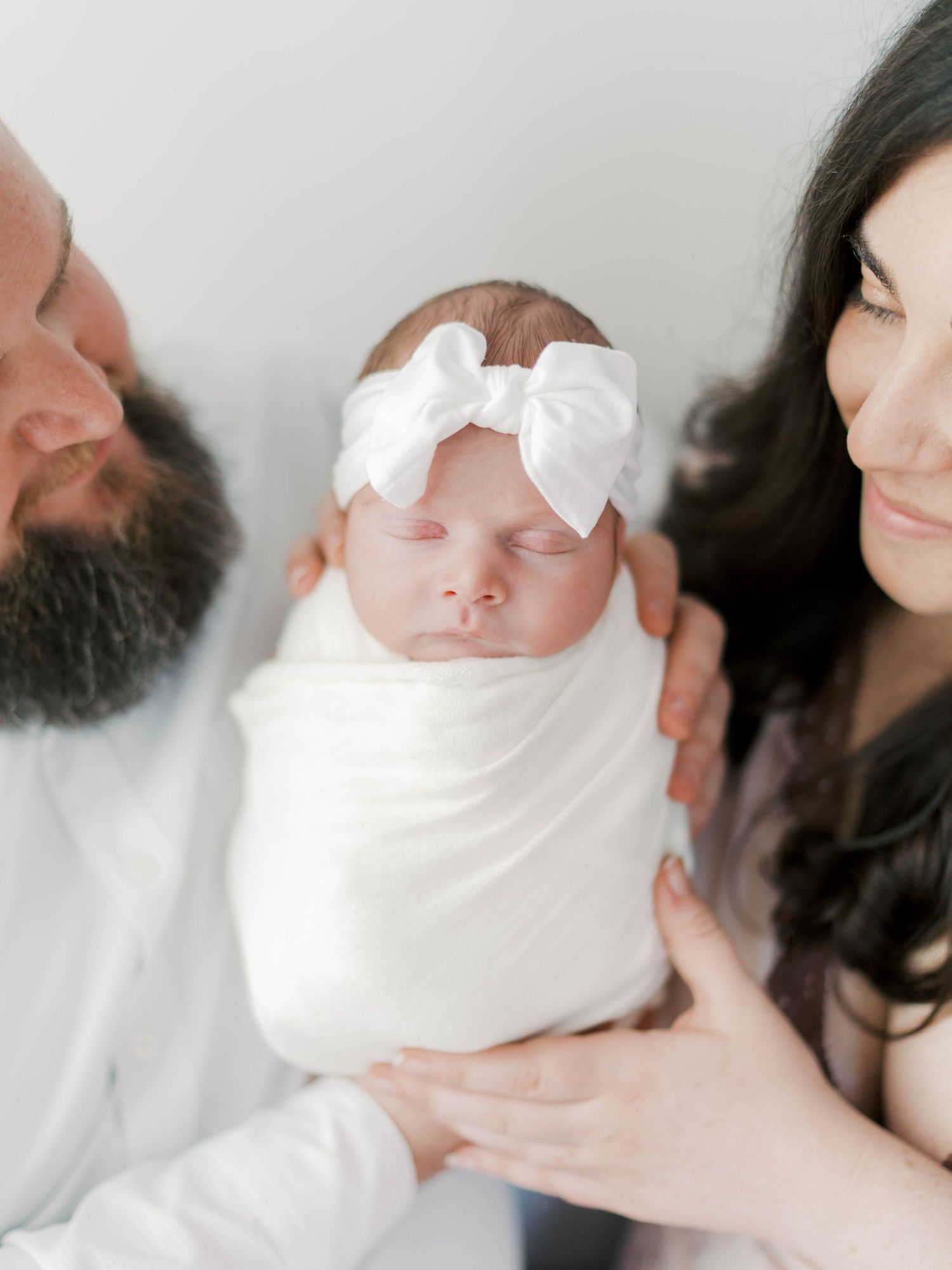 A bearded father and dark-haired mother lovingly hold their swaddled newborn baby girl wearing a white bow headband.