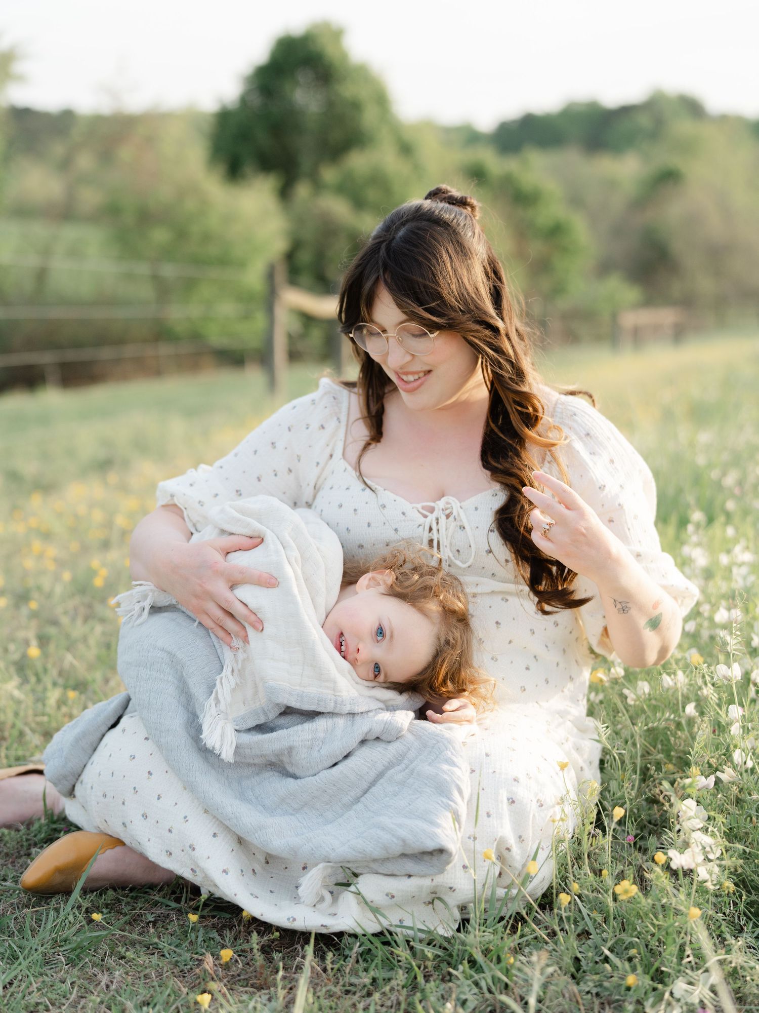 A mother with long brown hair sits in a sunny field, cradling her young child in her white floral dress.