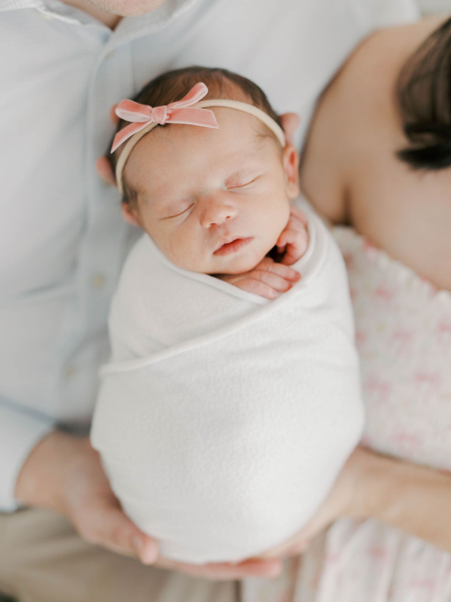A newborn baby swaddled in white fabric sleeps peacefully on a soft pink and white surface.