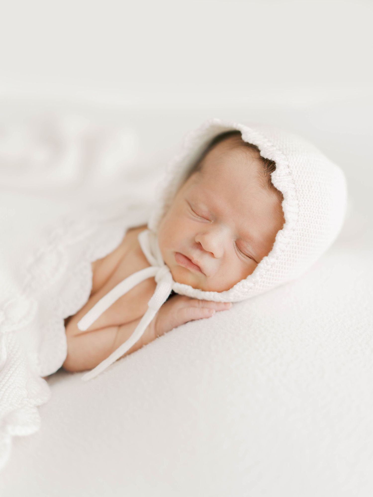 Sleeping newborn baby wearing white bonnet and wrapped in delicate white fabric.