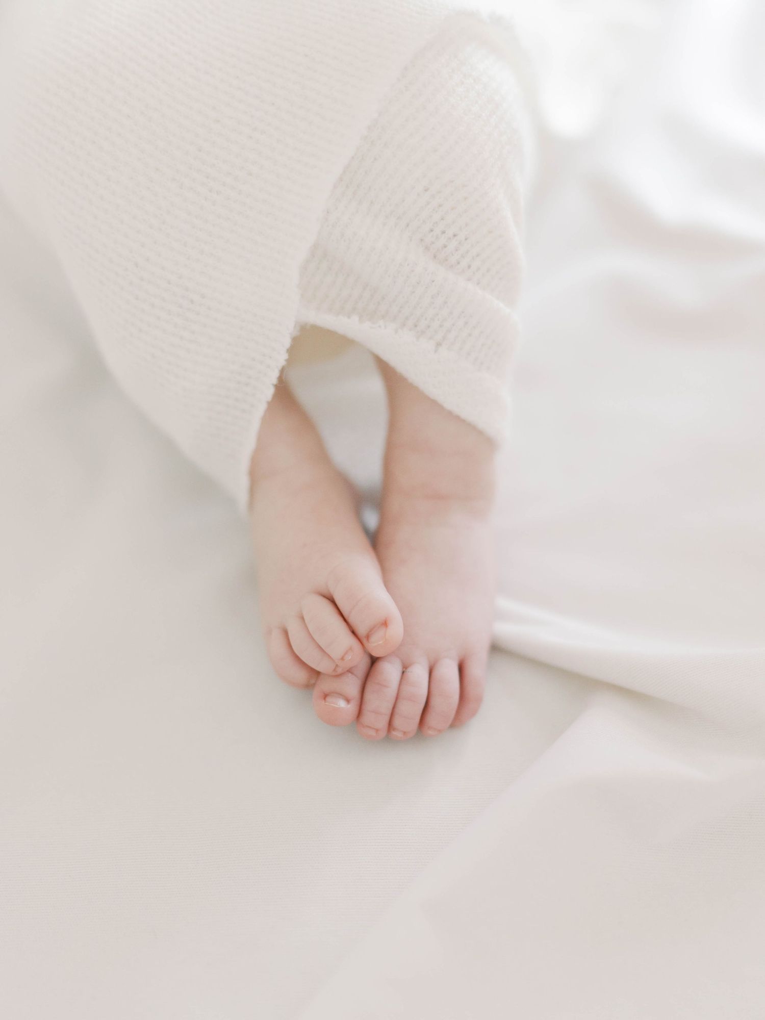 Tiny newborn baby feet peeking out from a soft white knit blanket on a white background.