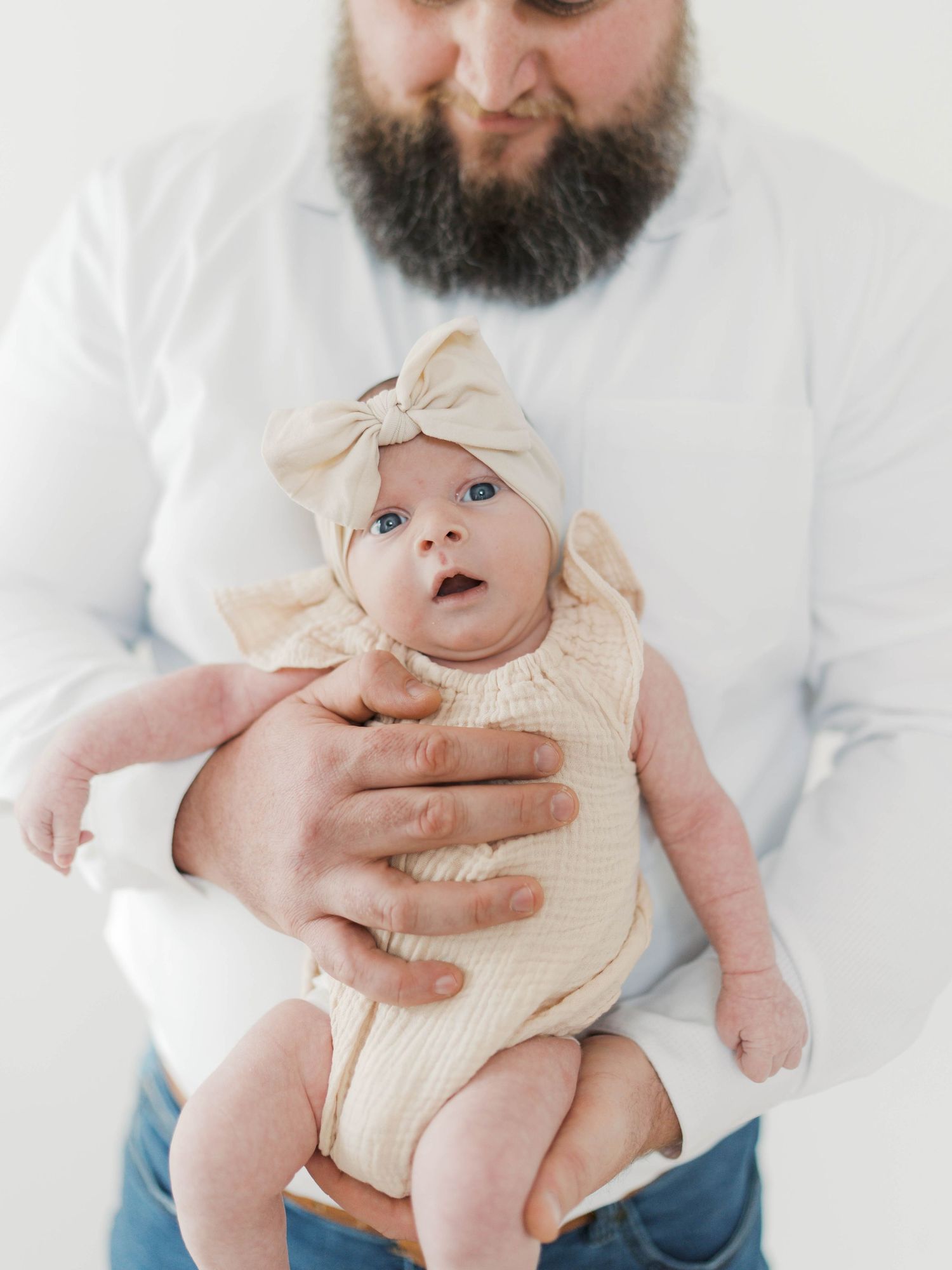 Bearded father in white shirt cradling newborn baby wearing cream romper and bow headband.