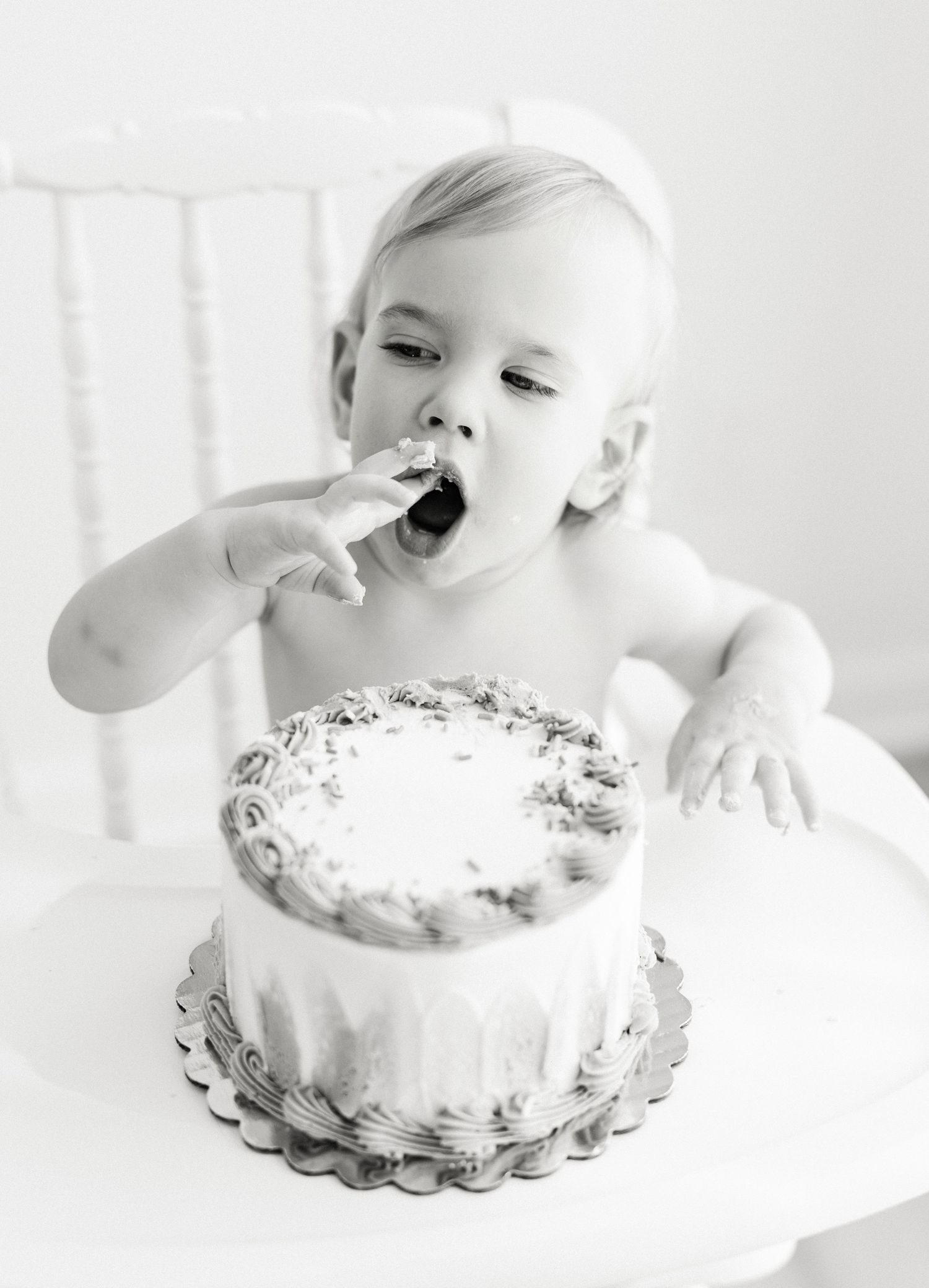 Black and white photo of a toddler in a high chair licking frosting from fingers while enjoying a decorated smash cake.