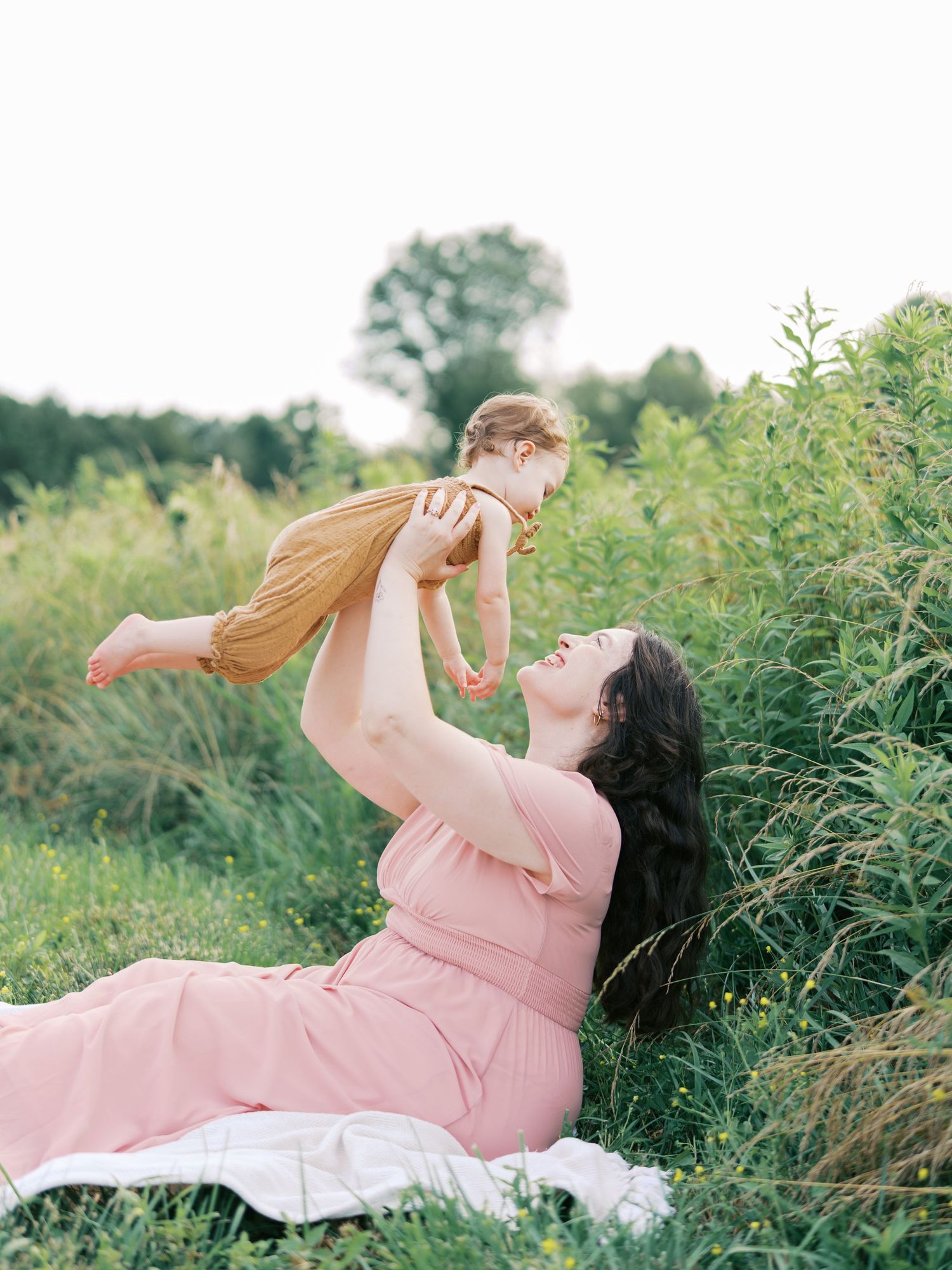 A person in a pink dress lies on a white blanket in a lush green field while playing with a baby.