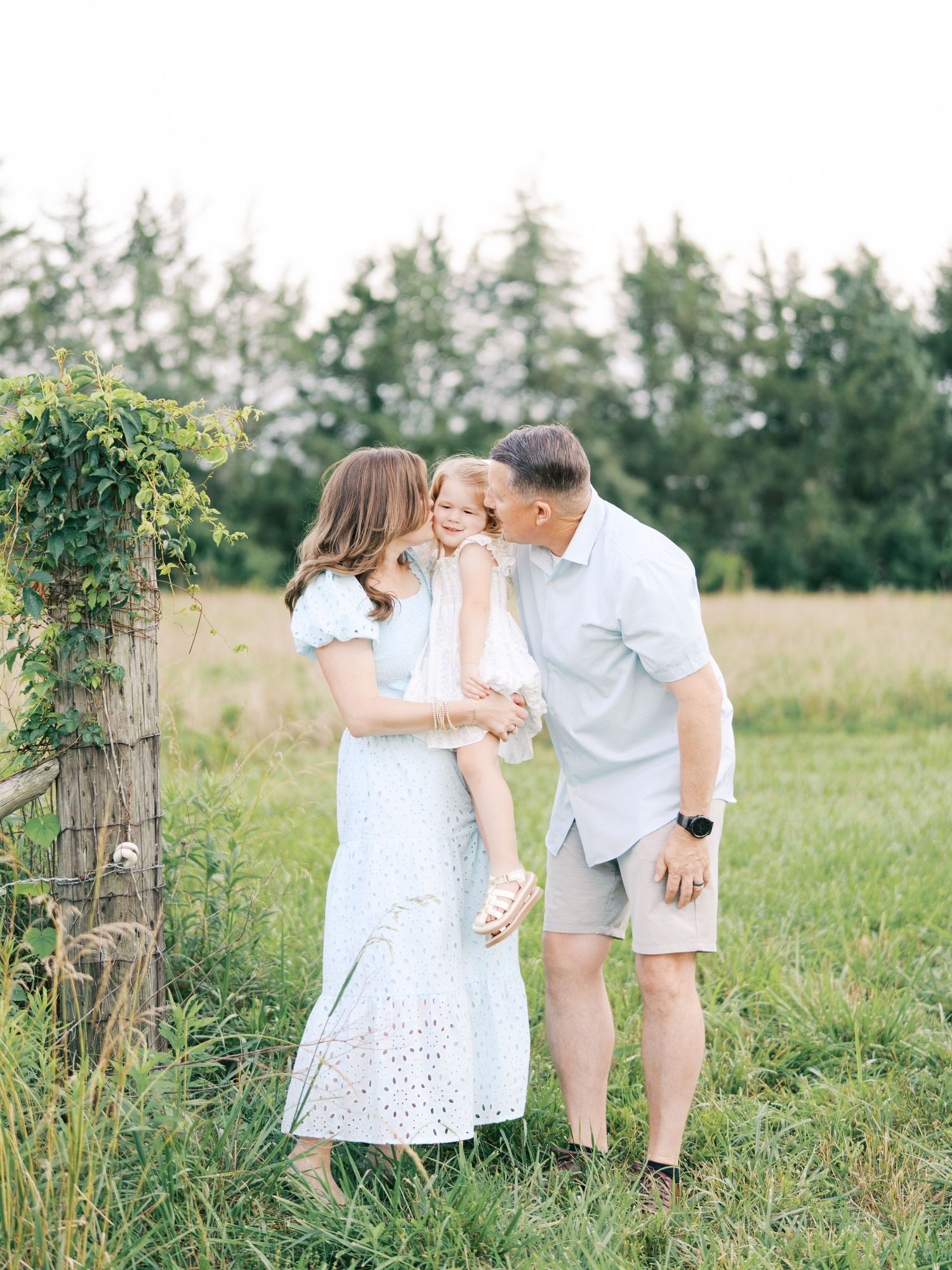 A couple shares tender moments in a sunny field with tall grass and trees in the background.