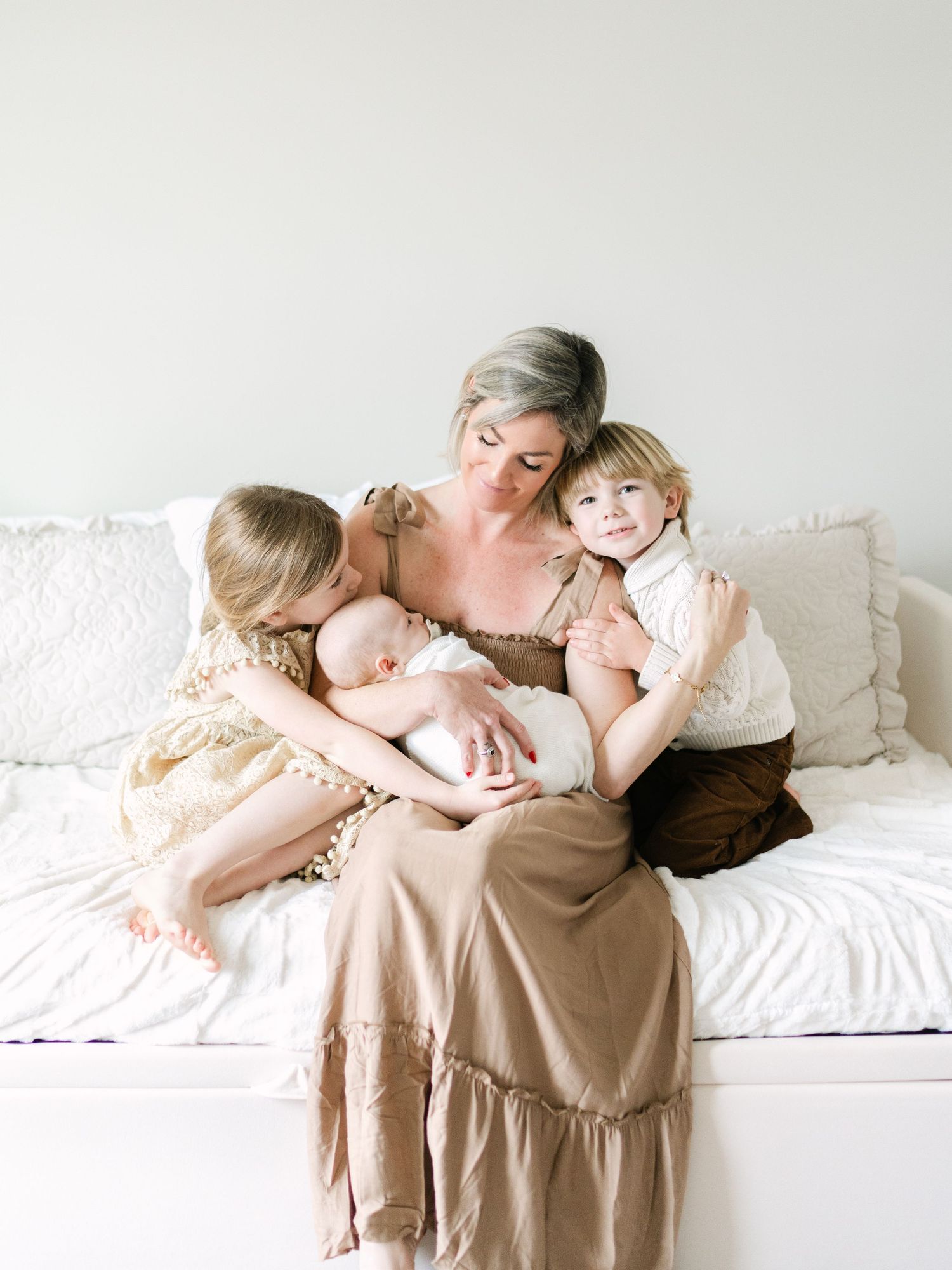A mother with gray hair sits on a white couch holding a newborn while two young children cuddle close in warm tones.