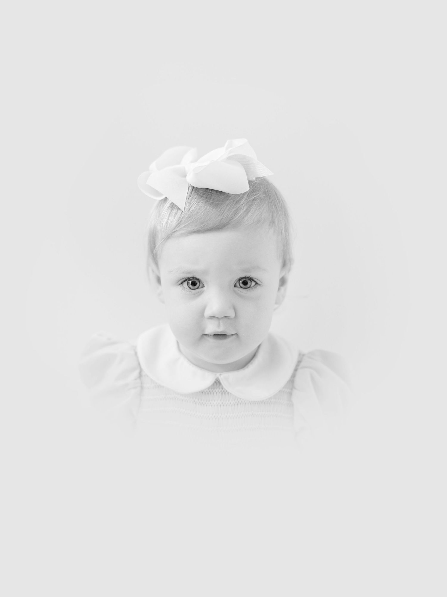 Black and white portrait of a baby girl wearing a white bow and peter pan collar dress.