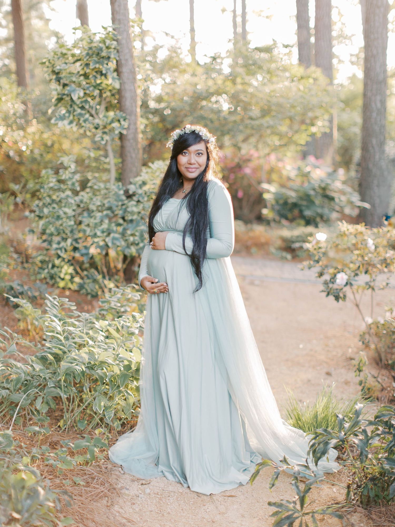 Pregnant woman in flowing blue dress and flower crown stands on forest path during sunset.