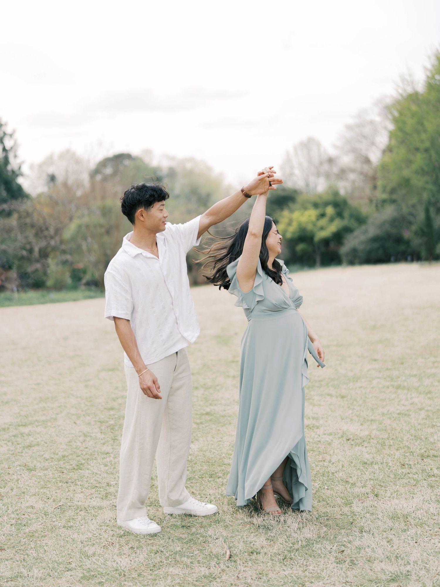 A couple dances in a grassy field during their maternity photoshoot wearing coordinated light-colored outfits.
