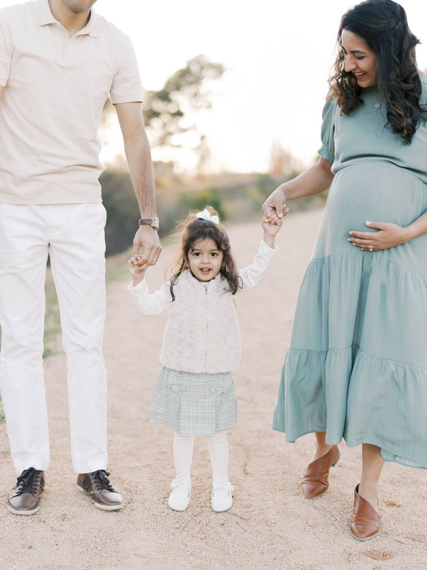 Pregnant woman in sage dress holds hands with toddler daughter and partner during outdoor maternity photo session.