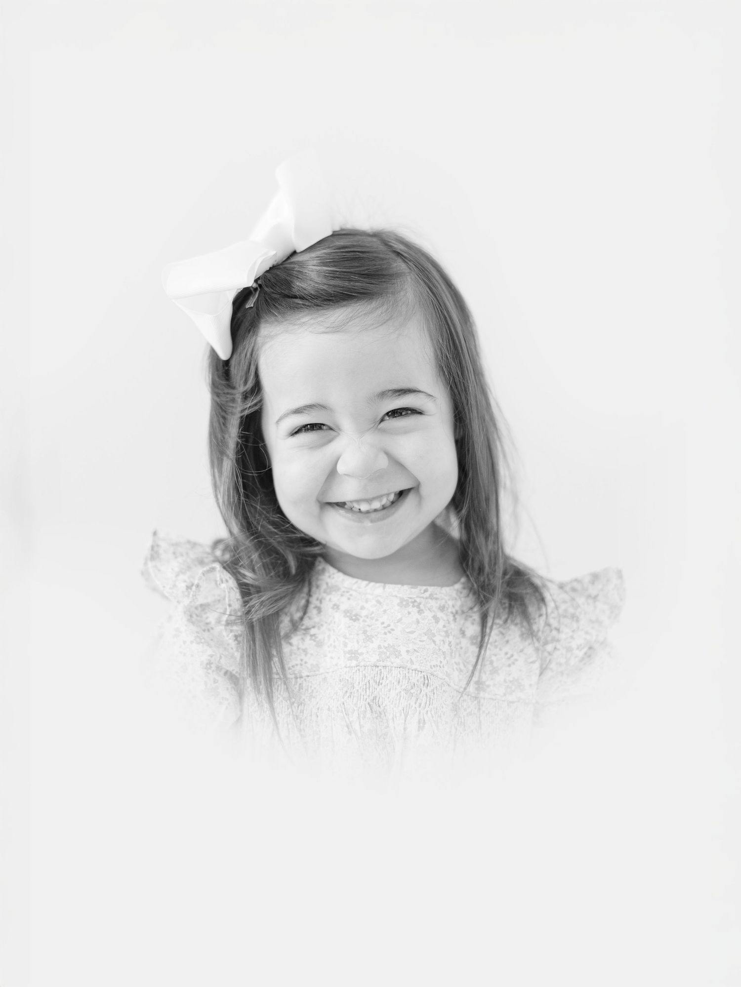 Black and white portrait of a smiling young girl with a white bow in her hair wearing a ruffled dress.