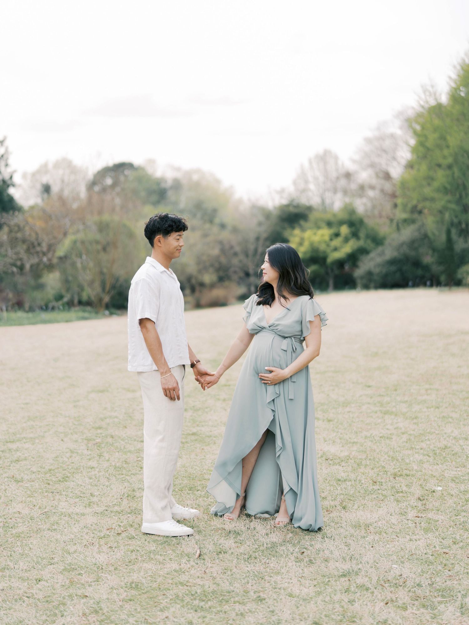 A couple stands in a grassy field wearing coordinated white and blue-gray outfits during a maternity photoshoot.