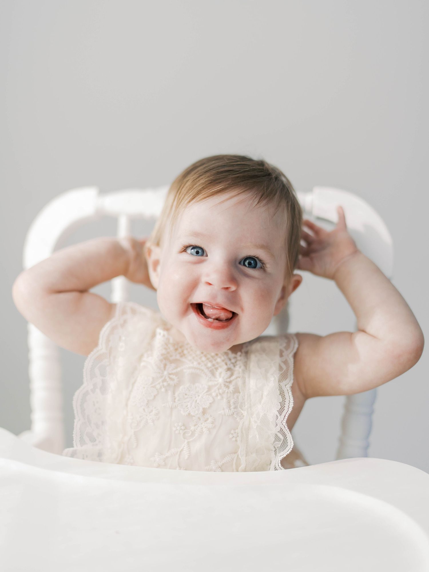 Joyful baby in lace dress laughing while sitting in white high chair.