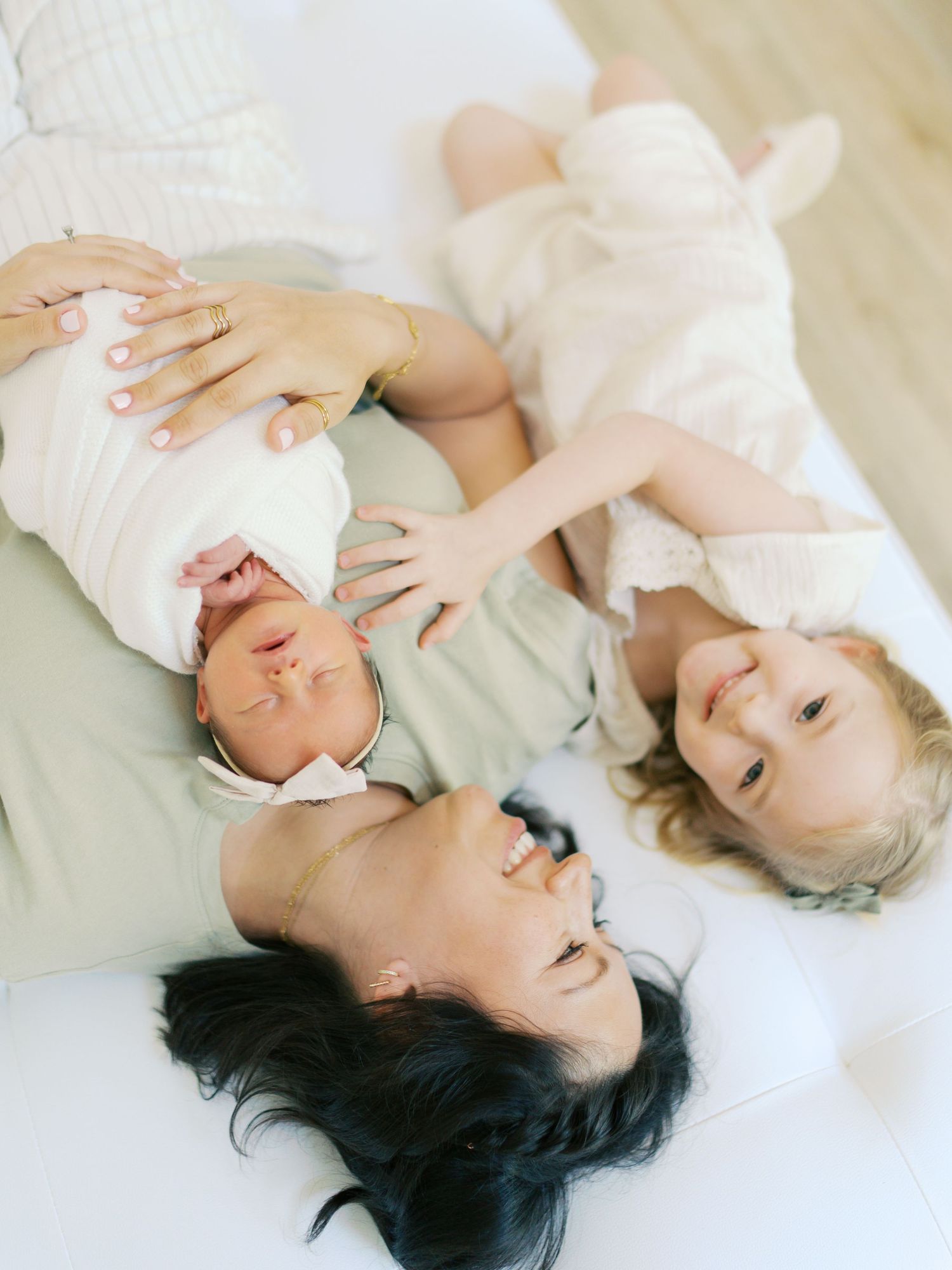 A mother lies on a white bed smiling, cradling a newborn in white swaddle, with a happy toddler girl beside her.