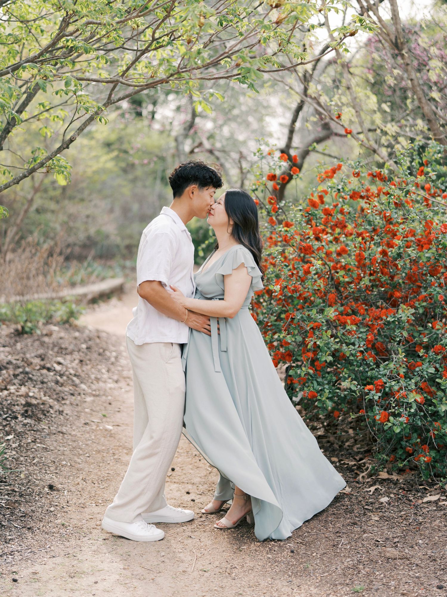 Romantic maternity portrait of couple by flowering bush on dirt path.