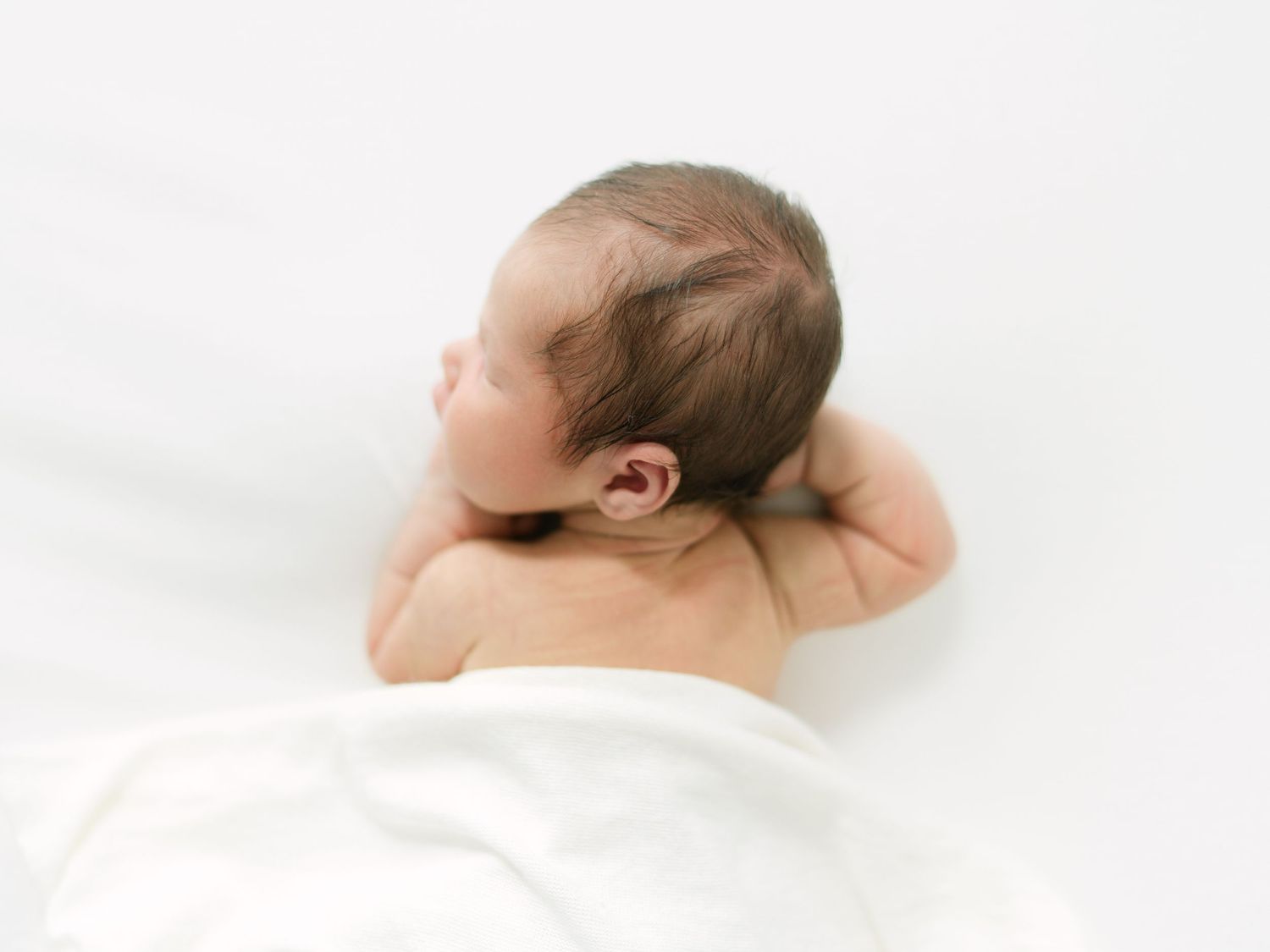 Newborn baby lying on white blanket, viewed from behind, showing tiny ears and dark hair.