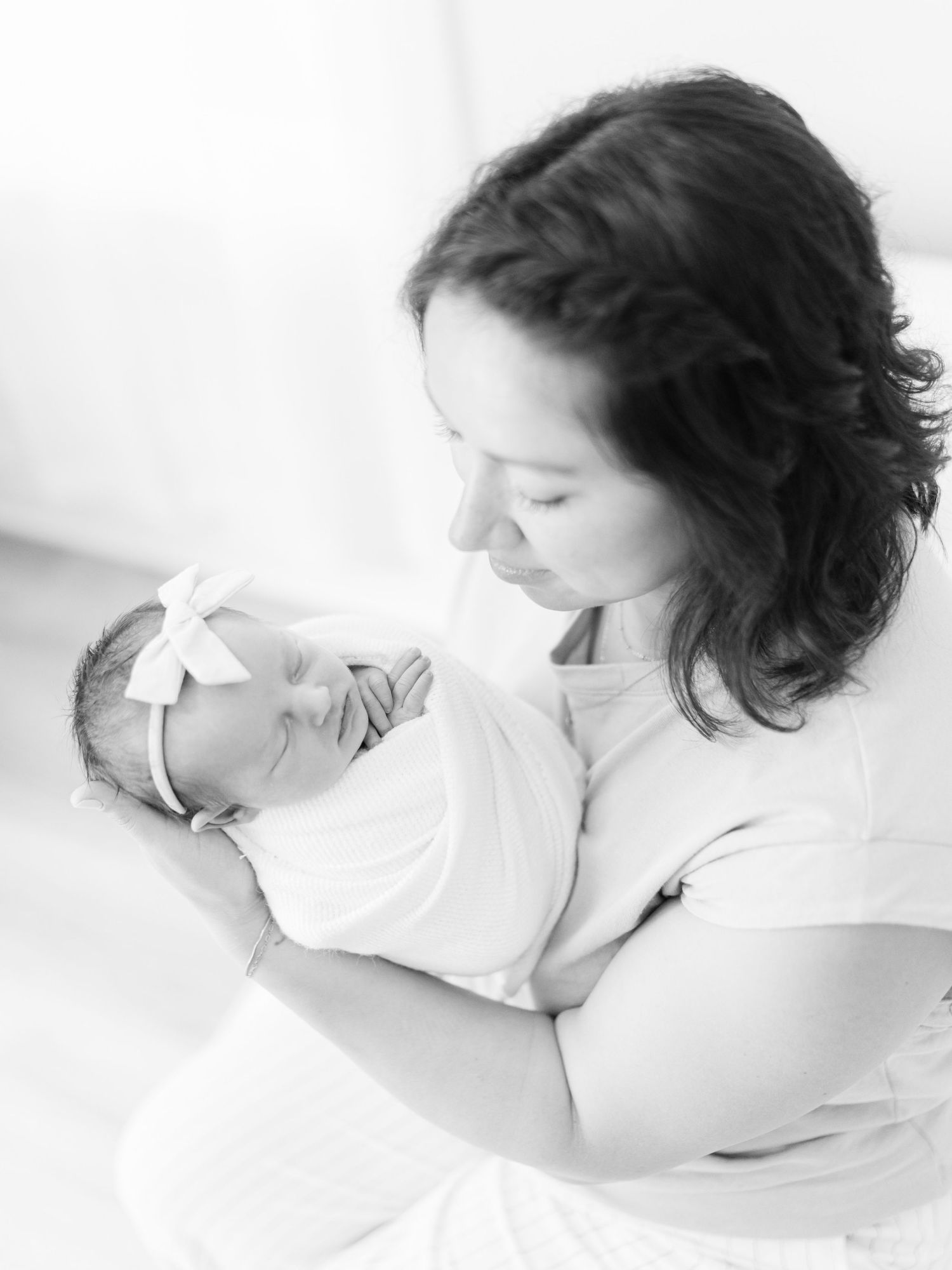 Black and white photo of a mother tenderly cradling her sleeping newborn baby girl wearing a white bow headband.
