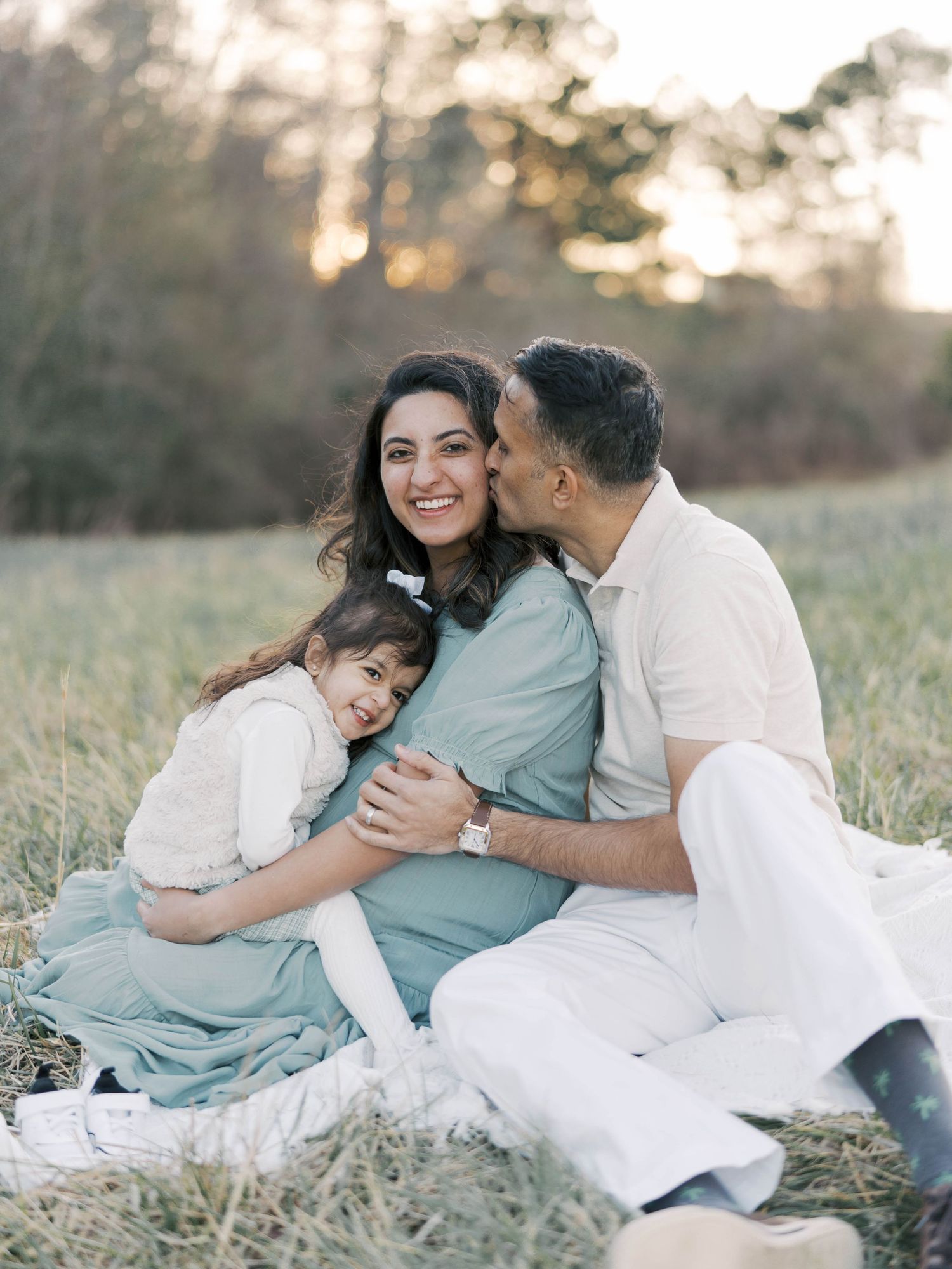Family cuddling together on blanket in grassy field at sunset.