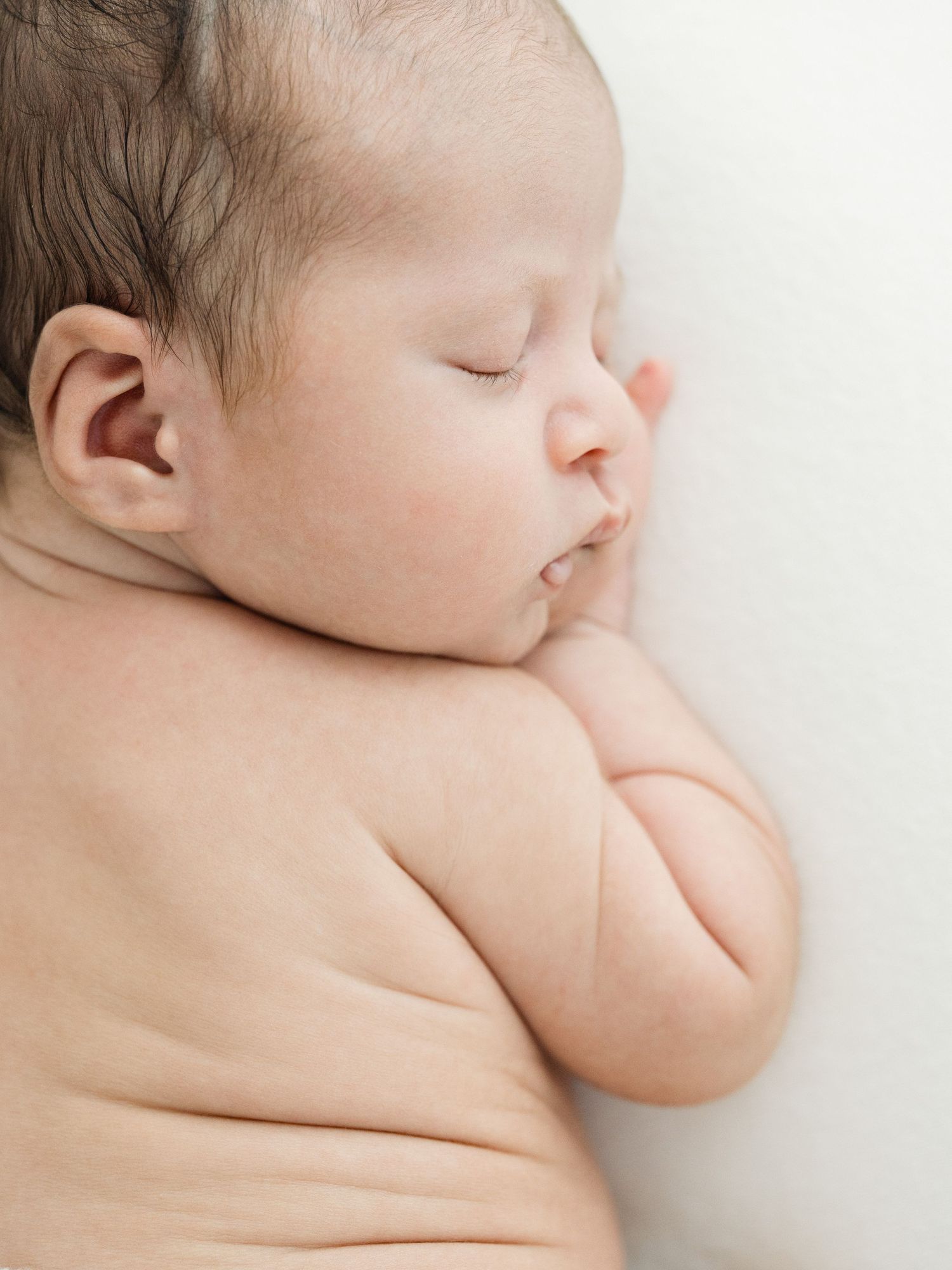Peaceful newborn baby sleeping on white background, curled up with chubby arms and dark hair after visiting lactation consultants in Raleigh