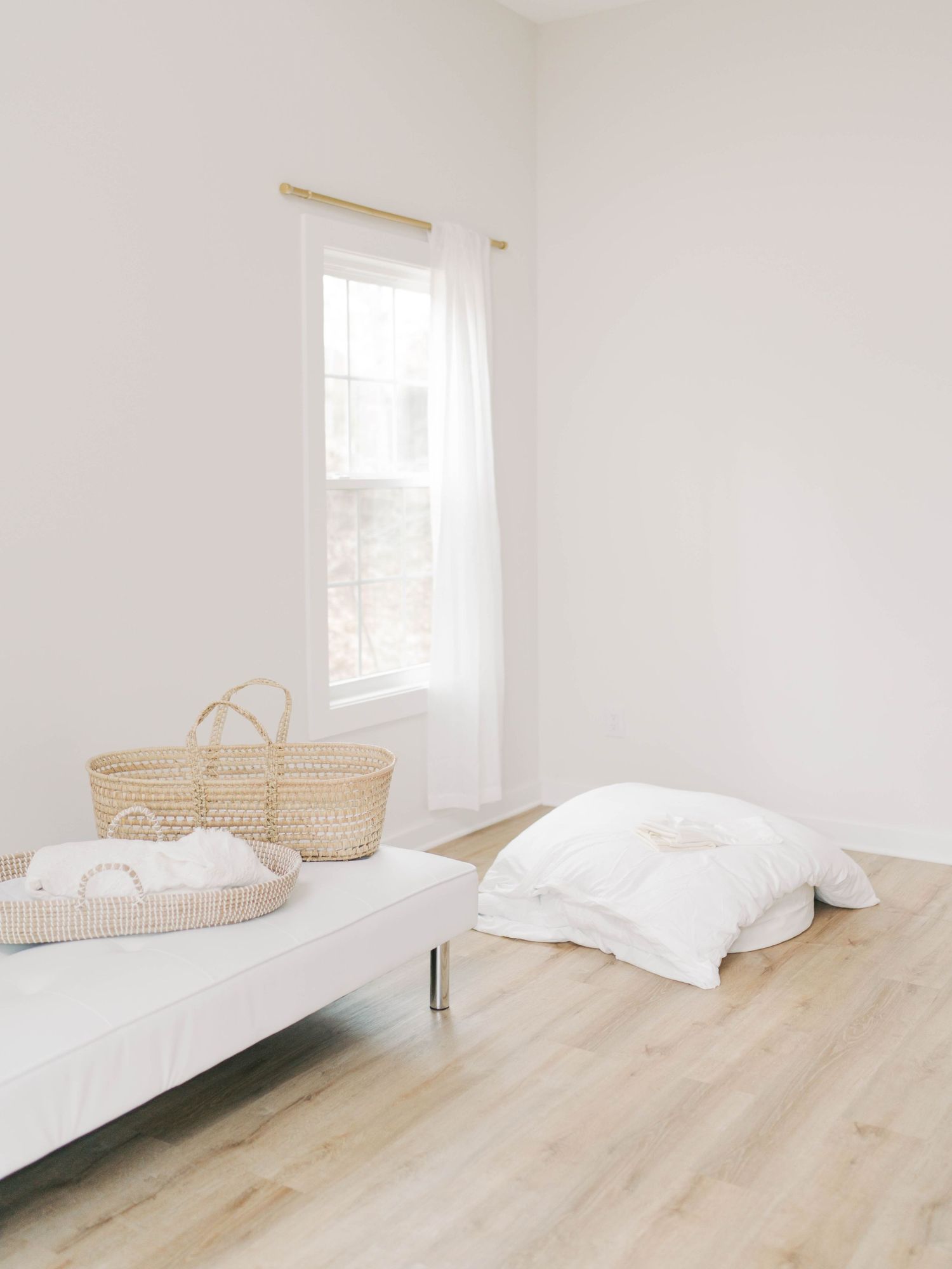 A minimalist white bedroom with natural light features a simple bed and woven baskets.