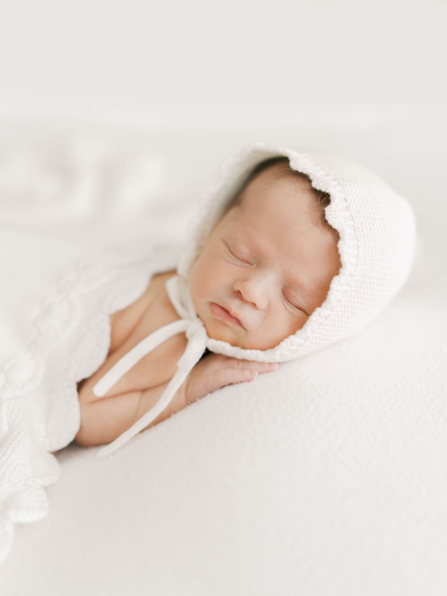 Sleeping newborn baby wearing a delicate white knit bonnet, swaddled in soft white fabric.