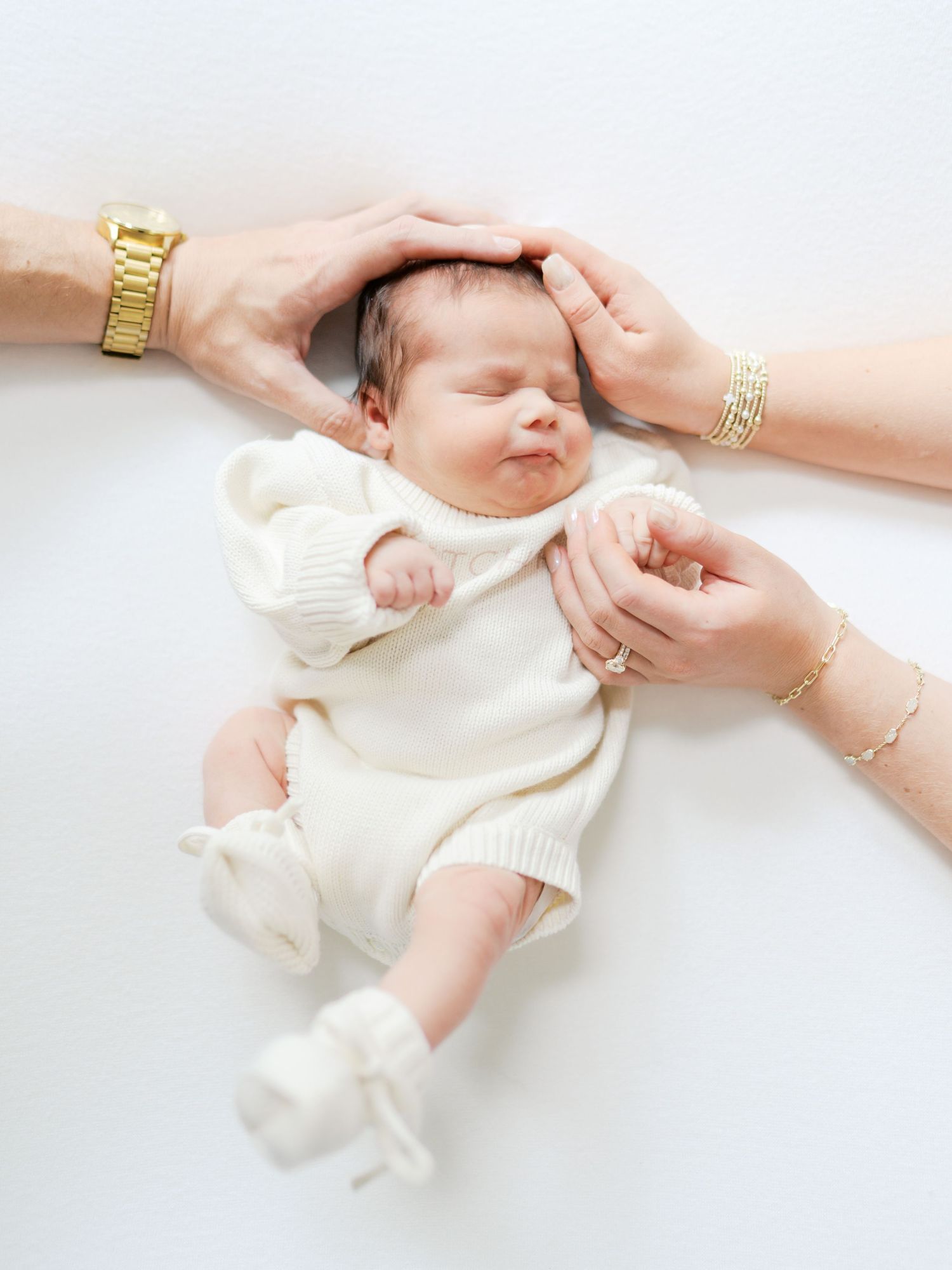 Newborn baby in white outfit lying peacefully as parents gently cradle head with loving hands.