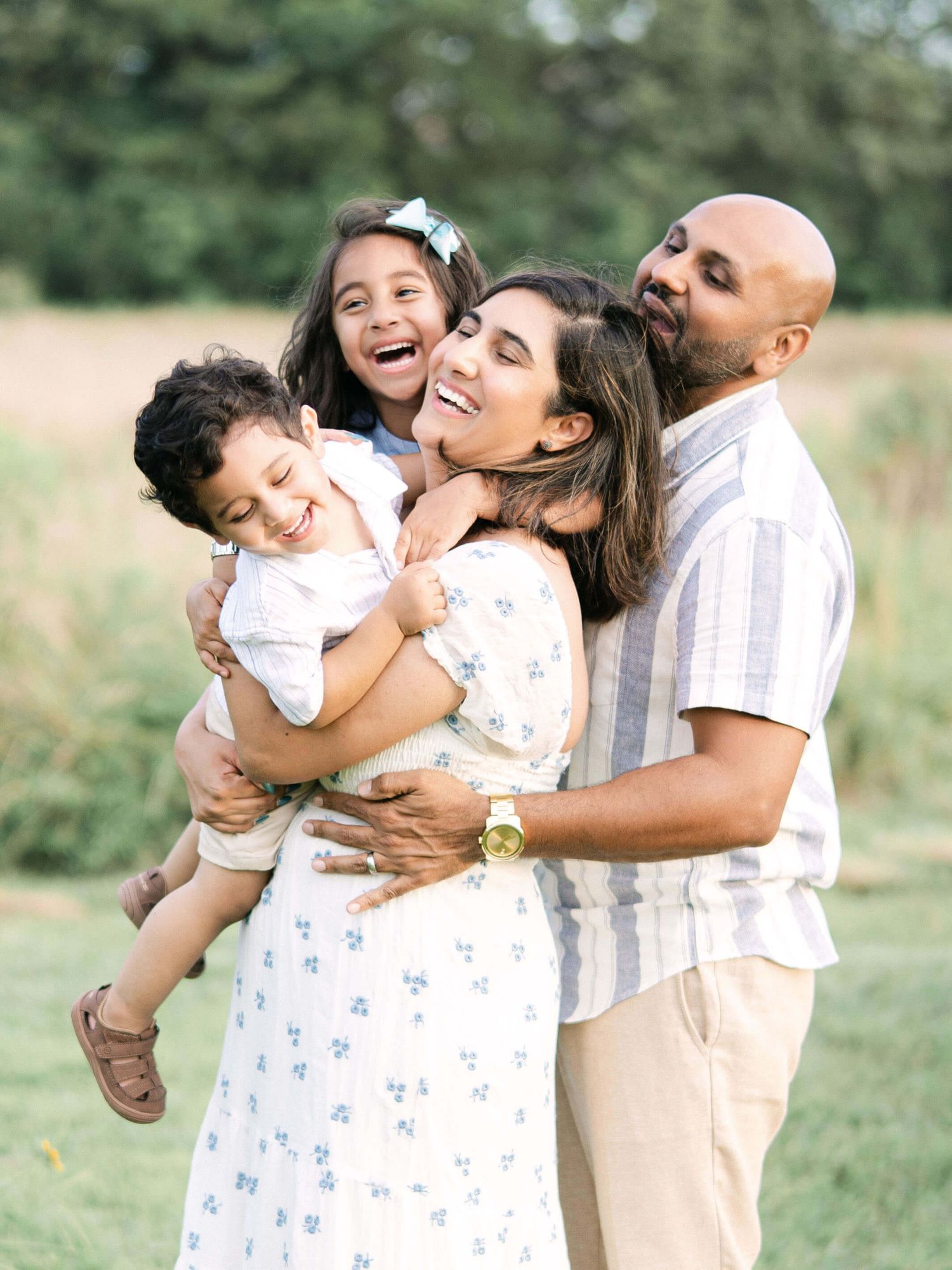 A joyful family of 4 shares a warm embrace in a sunlit field, with children laughing & smiling after visiting pediatric dentists in Raleigh