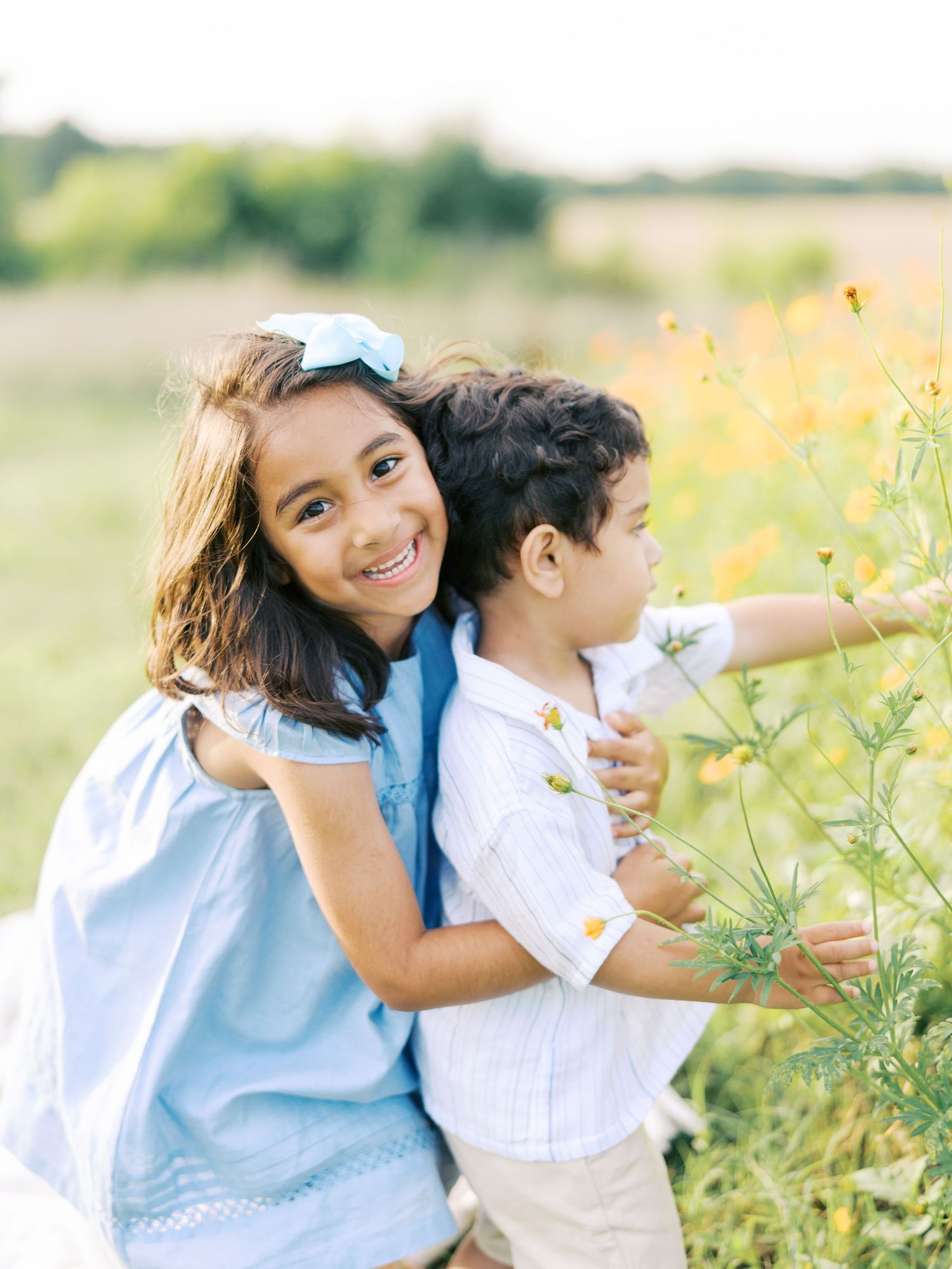 Two children in light blue and white outfits explore wildflowers together in a sunny meadow on a summer day.