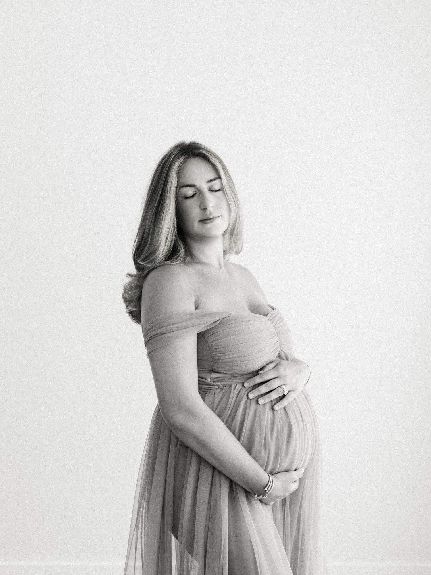 Black and white maternity photo of a woman in a flowing tulle gown cradling her baby bump after visiting midwives in Raleigh