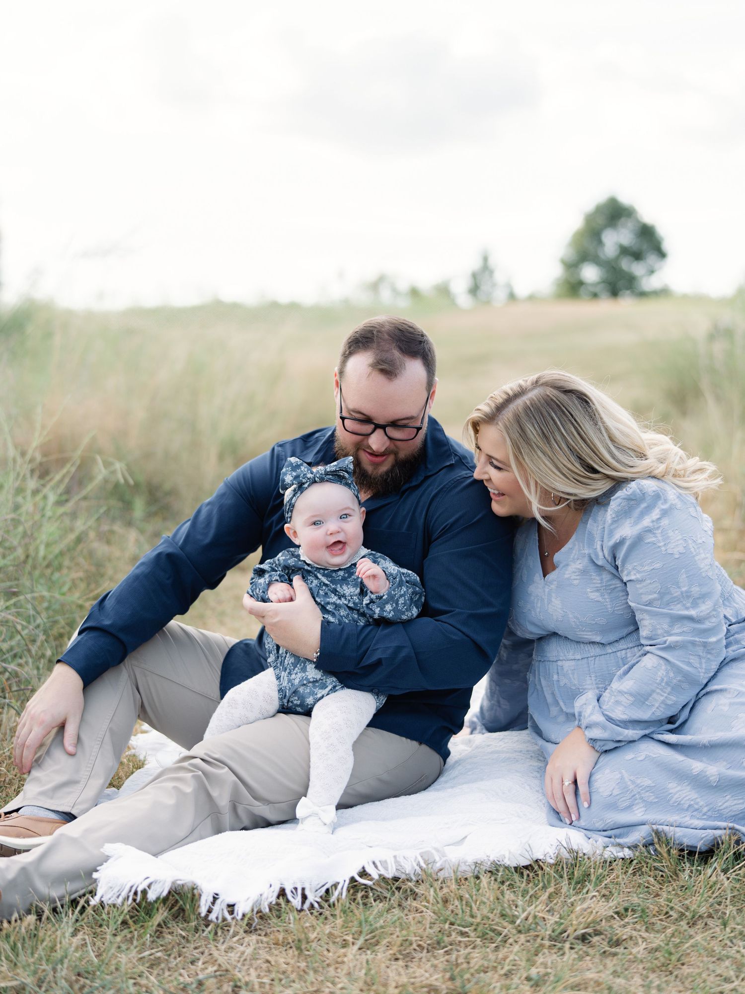 A young family sits on a white blanket in a field, parents in blue outfits smiling at their happy baby girl.