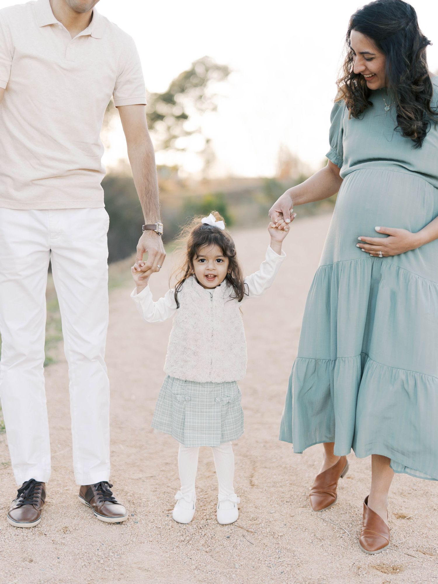 Family standing together on dirt path with pregnant woman in blue dress and small child holding hands between them.