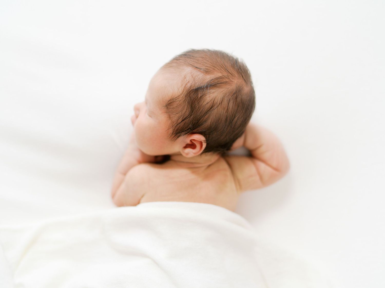 Newborn baby lying on white blanket, viewed from behind, with tiny hands tucked under head.