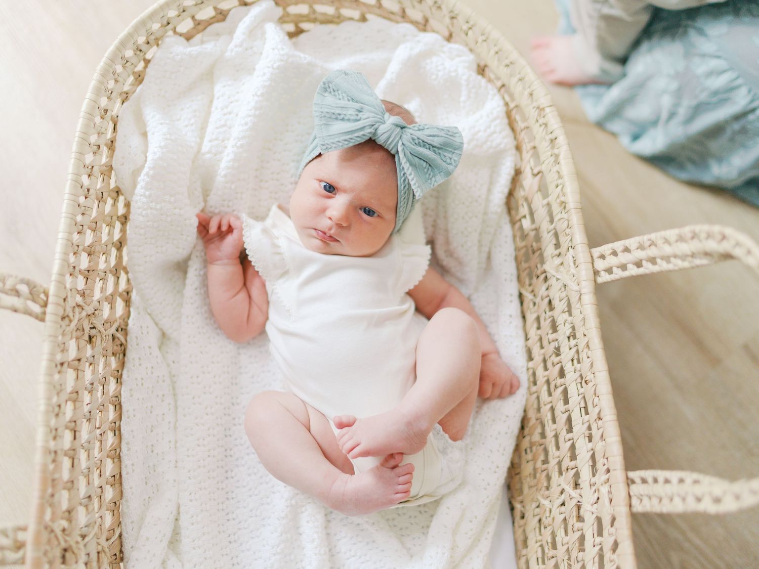 Newborn baby in white onesie and sage green bow headband lying in a wicker Moses basket with white blanket.