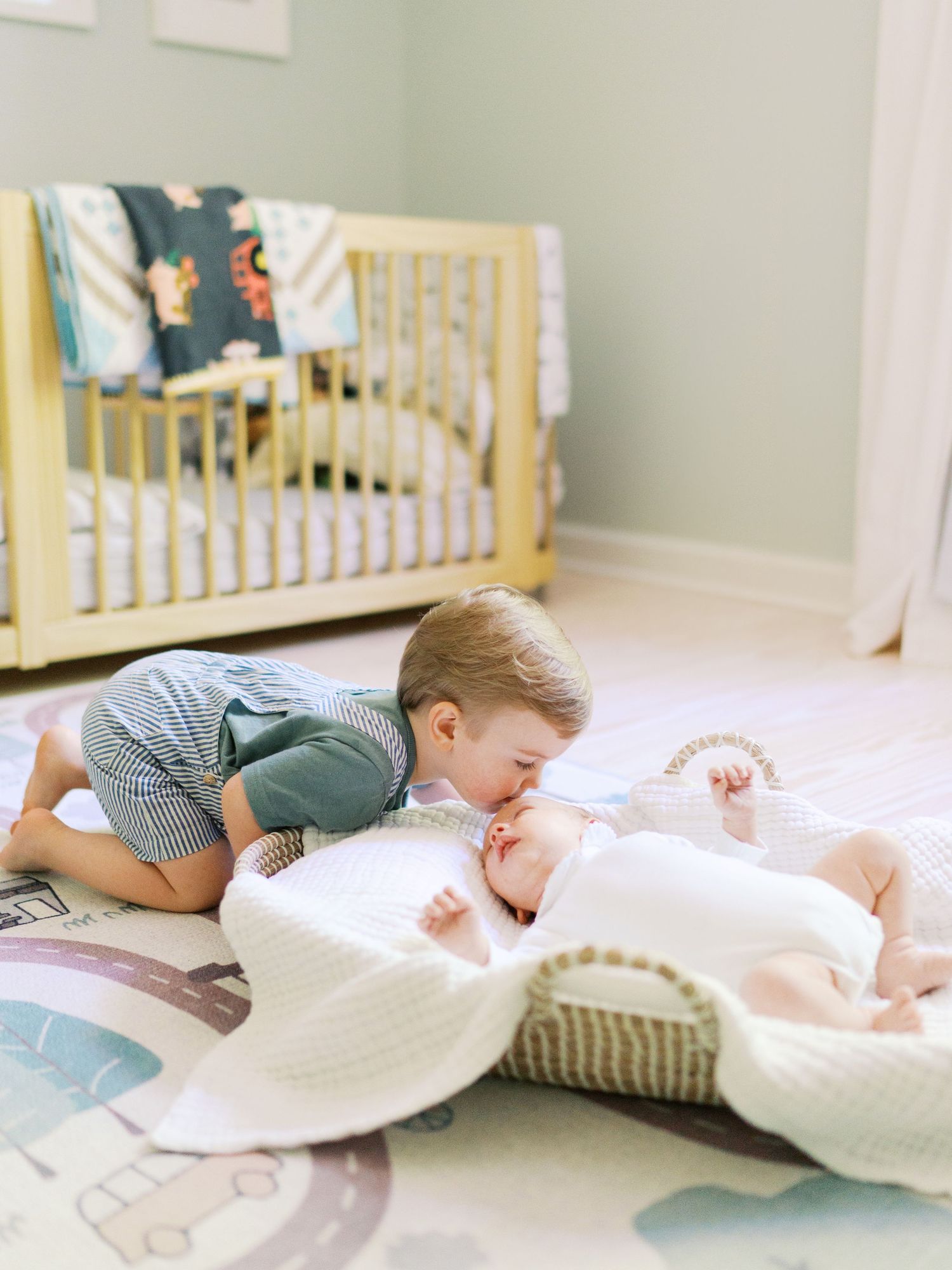 Toddler boy gently kissing newborn baby sibling lying in Moses basket on nursery floor near yellow crib.