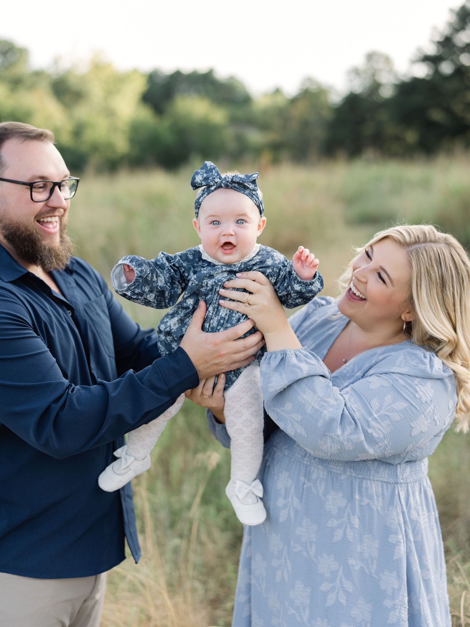A happy family of 3, with parents lifting their smiling baby girl outdoors in a sunny green field after a prenatal fitness class in Raleigh