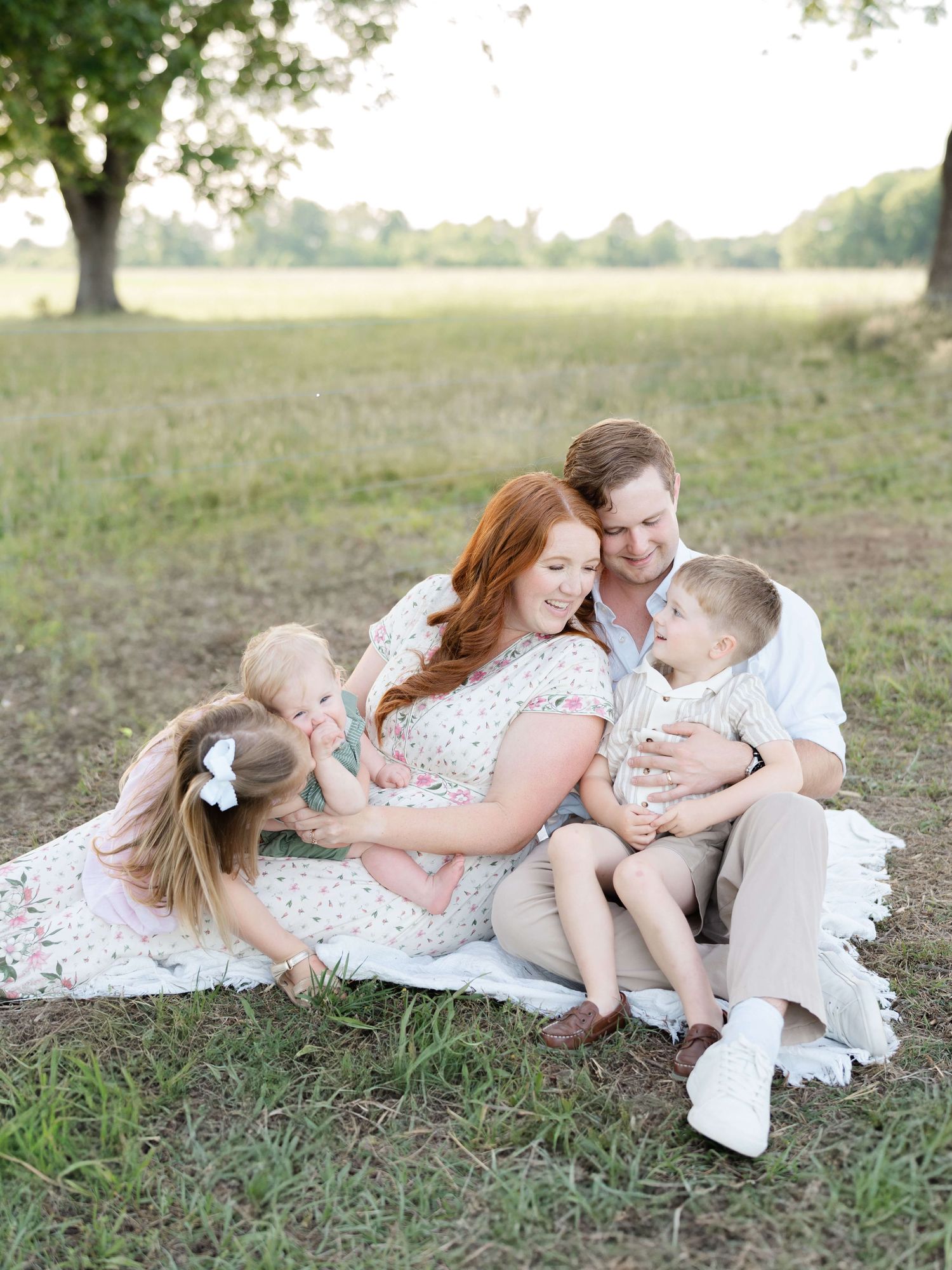 A pregnant woman with red hair sits on a blanket in a field with her husband and two young children.