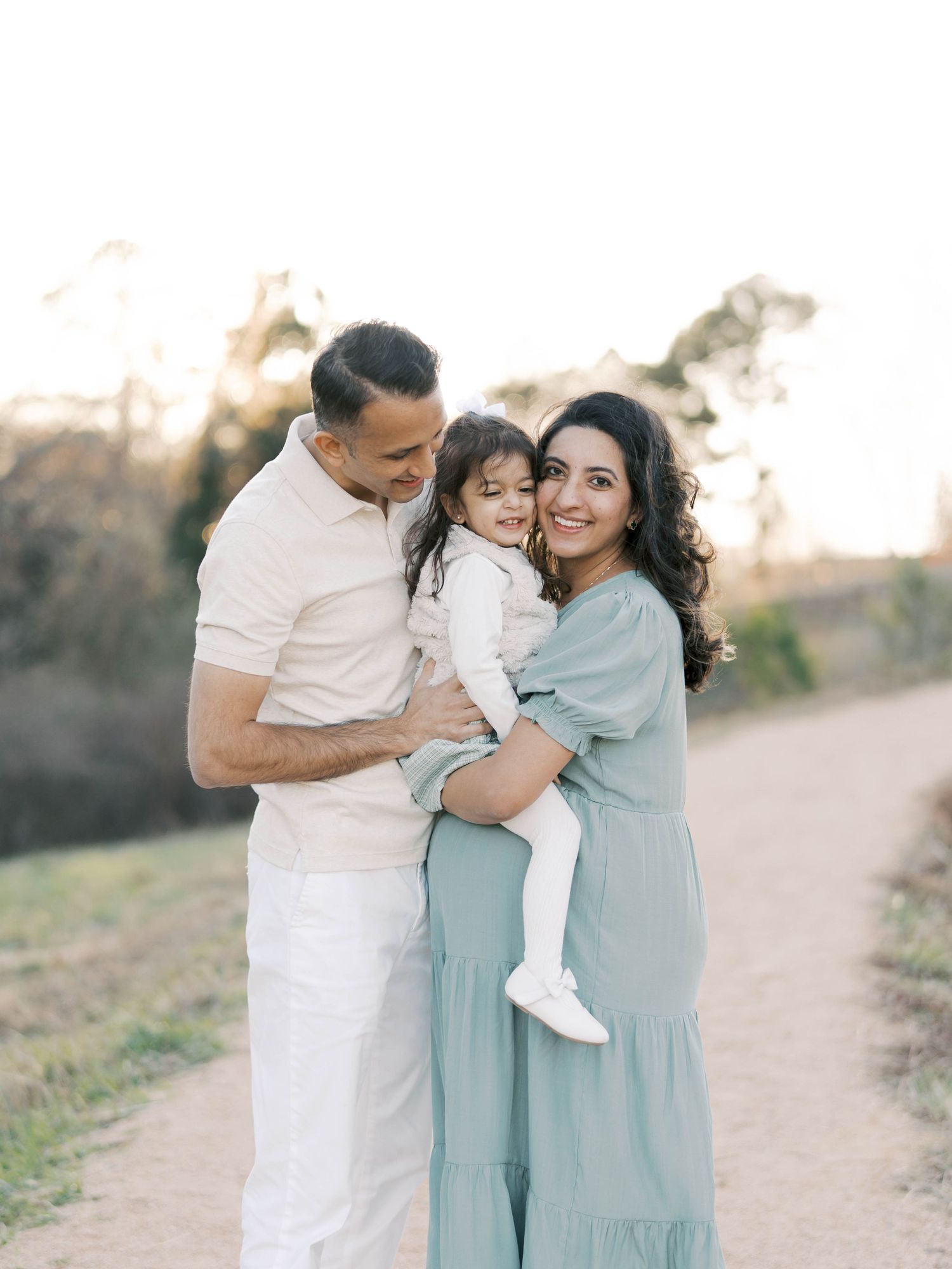 Family of three embracing outdoors in coordinated white and blue outfits at sunset.
