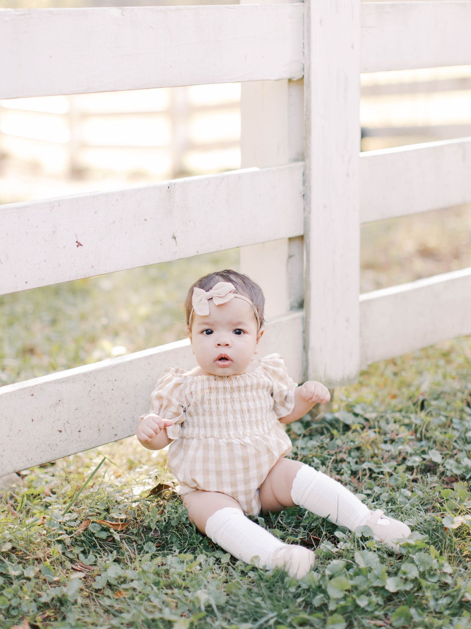 Baby in striped dress and white socks sitting by white fence in garden setting.