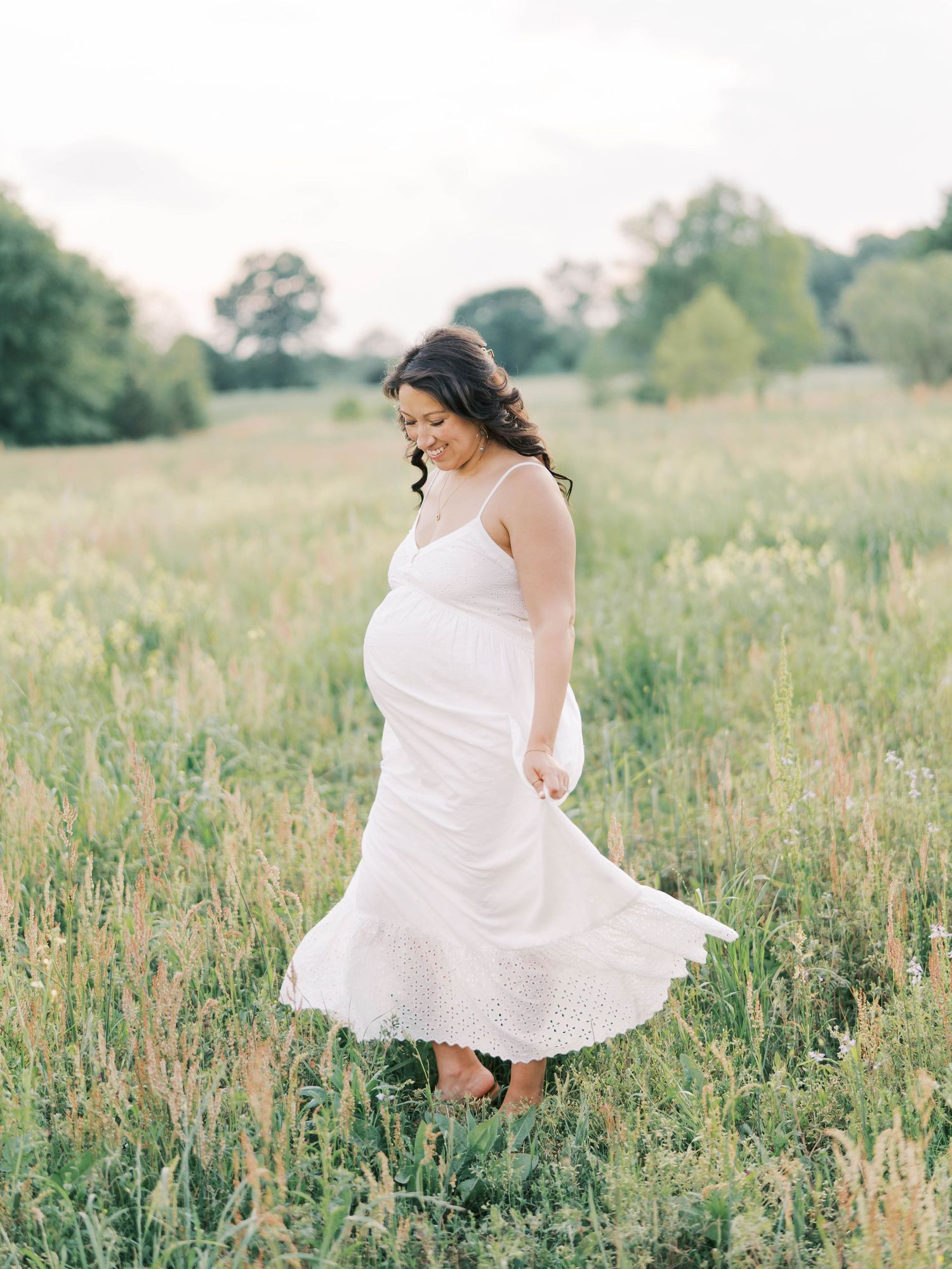 Pregnant woman in white sundress twirling in a lush green meadow during golden hour maternity photoshoot.