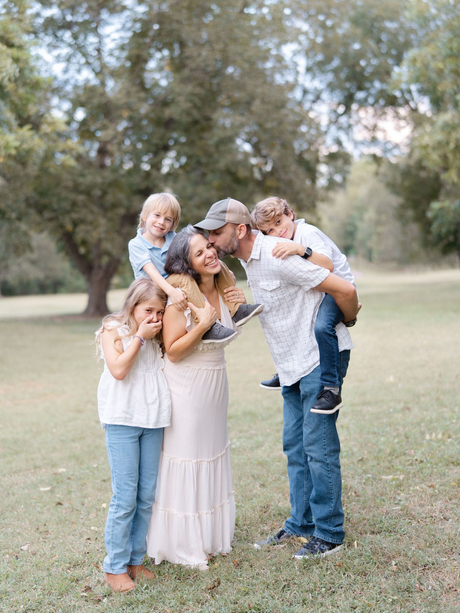 A joyful family of five laughs together outdoors in a park, with parents and three young children in casual neutral outfits.