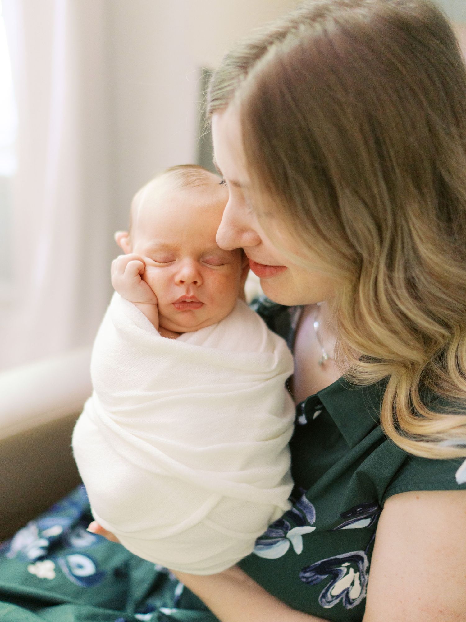 A mother tenderly holds her swaddled sleeping newborn baby close to her face in a soft, natural light setting.