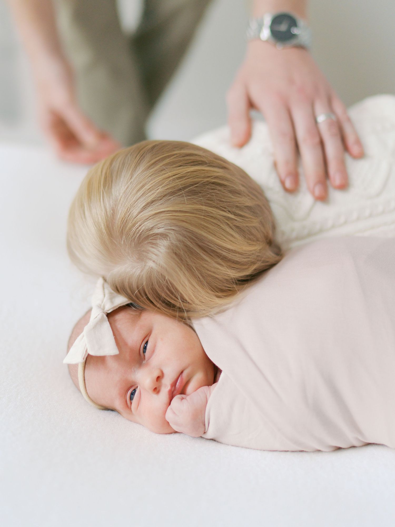 Newborn baby swaddled in pink wrap with bow headband lying on white bed during professional photo session.