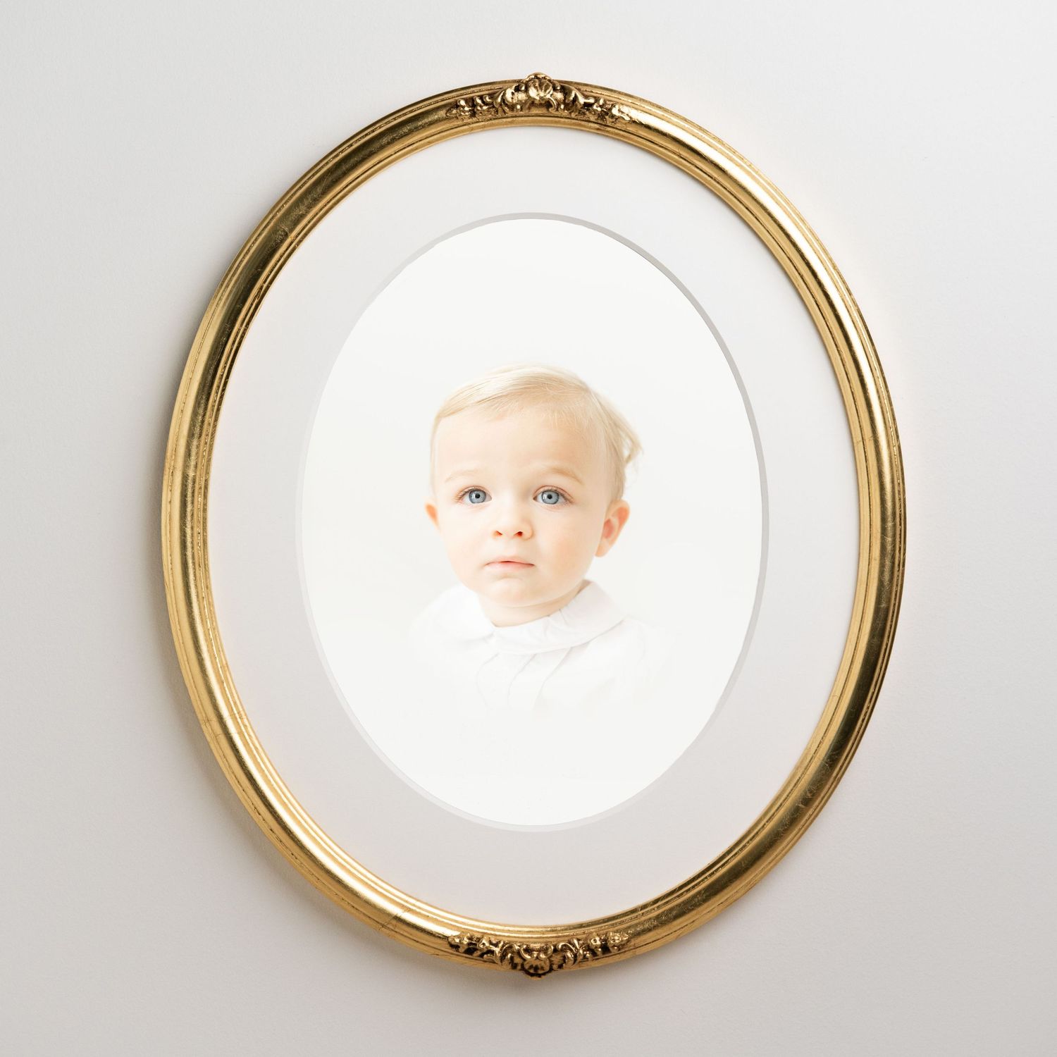 A baby portrait displayed in an ornate oval gold frame with white matting against a gray wall.